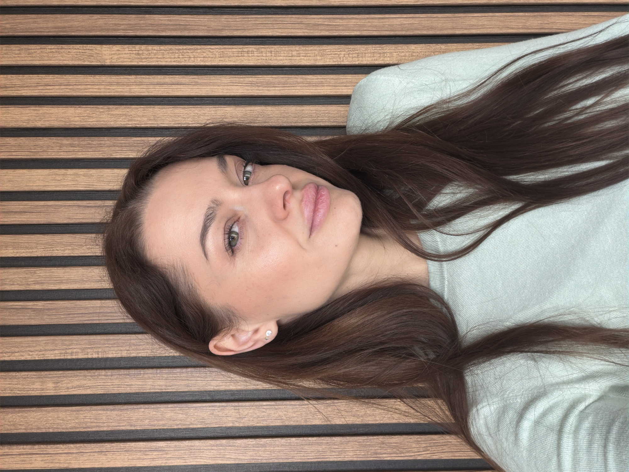 A woman with brown hair and green eyes lies on a wood-paneled surface. She wears a light green sweater. Her face is tilted towards the camera. The background consists of dark and light brown parallel panels. The location cannot be identified based on the image.