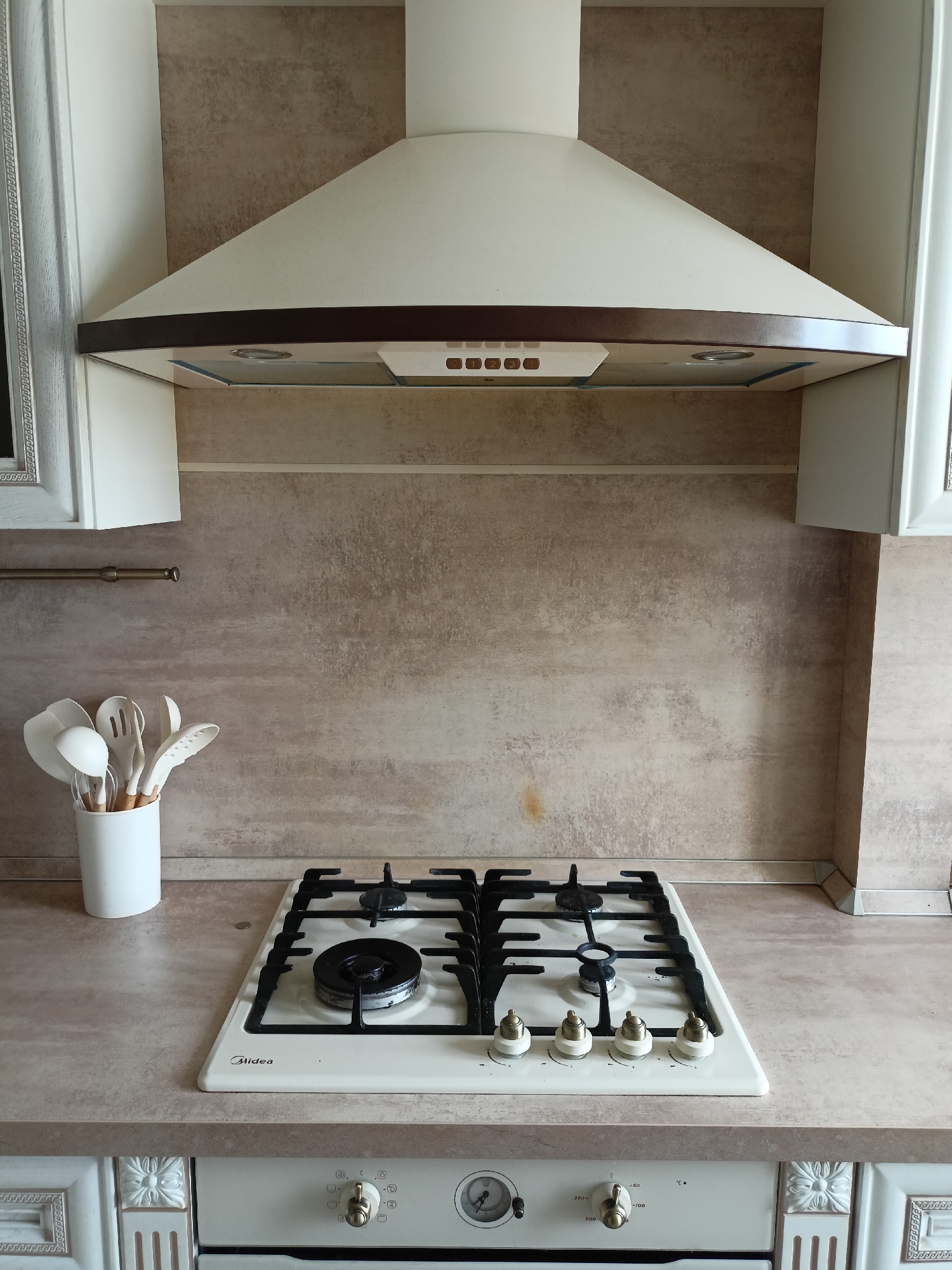 The image shows a clean kitchen countertop with a white gas stove and a white range hood. On the left side of the countertop, a white ceramic container holds several white kitchen utensils, including spatulas and slotted spoons. The backsplash behind the stove and utensils is a textured, light brown material. The range hood is white with a dark brown trim and appears to be a standard model. The stove itself is white with black grates and burners. The control knobs for the stove are visible, as well as the "Midea" brand name. Below the countertop, part of a white oven is visible, with a control dial and a temperature gauge. The overall scene suggests a well-maintained kitchen. There is no activity taking place, and the lighting suggests it is daytime. The location context provided is Volgodonsk, Russia.