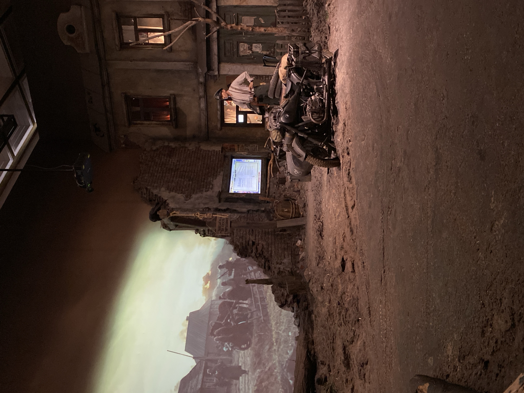 The image depicts a diorama or museum exhibit, likely relating to World War II or a similar conflict, set in a stylized urban environment.
Buildings: The backdrop features detailed facades of buildings, possibly representing a European city, with visible windows, brickwork, and signs of damage or age.
Vehicles: A motorcycle with a sidecar is prominently displayed in front of the building, possibly German in origin, suggesting a military presence.
People: A mannequin dressed in a military uniform stands next to the motorcycle, further reinforcing the military theme. There is also a large backdrop/ mural of figures with weaponry, possibly a battlefield scene.
Environment: The diorama includes elements to simulate a war-torn street, such as rubble, uneven ground, and debris.
Technology: A flat-screen display or monitor is visible within the diorama, likely providing additional information or context to the exhibit.
Based on the diorama’s content, particularly the motorcycle, mannequin, and backdrop, it is plausible that this is an exhibit related to World War II or a similar historical event, and it may be on display at the Moscow Victory Museum or a similar museum.