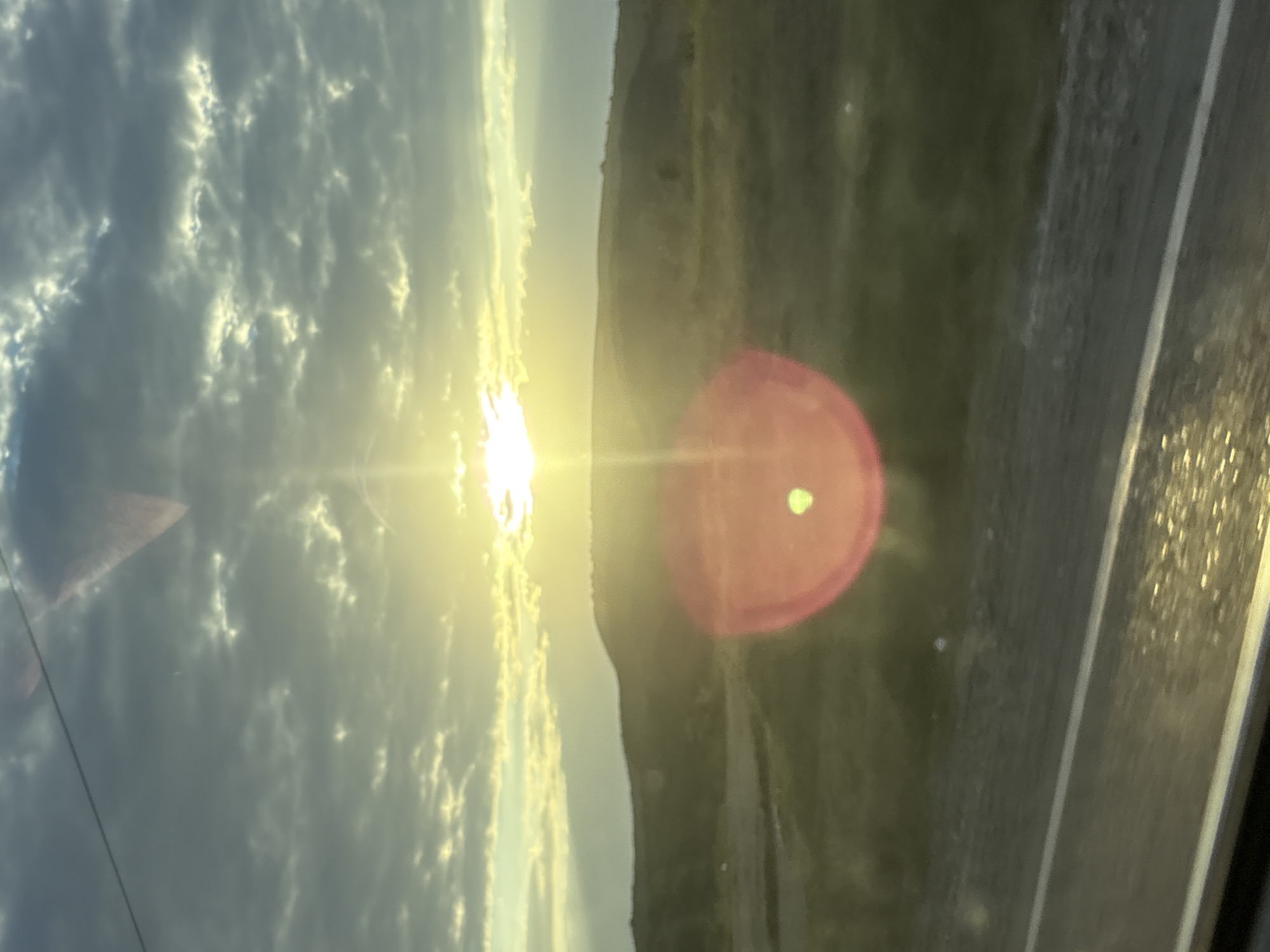 The image depicts a sunset over a rural landscape, likely near the village of Lysogorskaya, Russia. The sun is low on the horizon, partially obscured by clouds, casting a golden light over rolling hills covered in green grass. A prominent lens flare, a bright red circle with a green spot, is visible in the foreground, indicating the presence of a light source affecting the camera. The sky is a mixture of blue and gray clouds, with patches of sunlight breaking through. A road or path runs along the bottom of the image, with what appears to be a white line marking its edge. The overall impression is one of natural beauty and tranquility, captured during the transition from day to night.