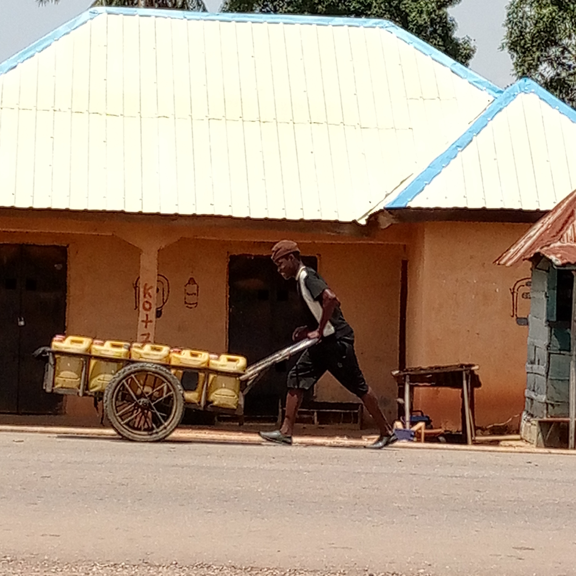 A man is pulling a cart loaded with yellow jerrycans along a paved road in Mararaba, Nigeria. The man is wearing a brown cap, a short-sleeved black and white shirt, dark shorts, and dark shoes. The cart has a single axle and large, spoked wheels. Behind the man is a low, single-story building with a light yellow roof and light blue trim. There are drawings on the wall of the building. To the right of the building is a small, dilapidated structure. A small table is also next to the building. The road is cracked and shows signs of wear.