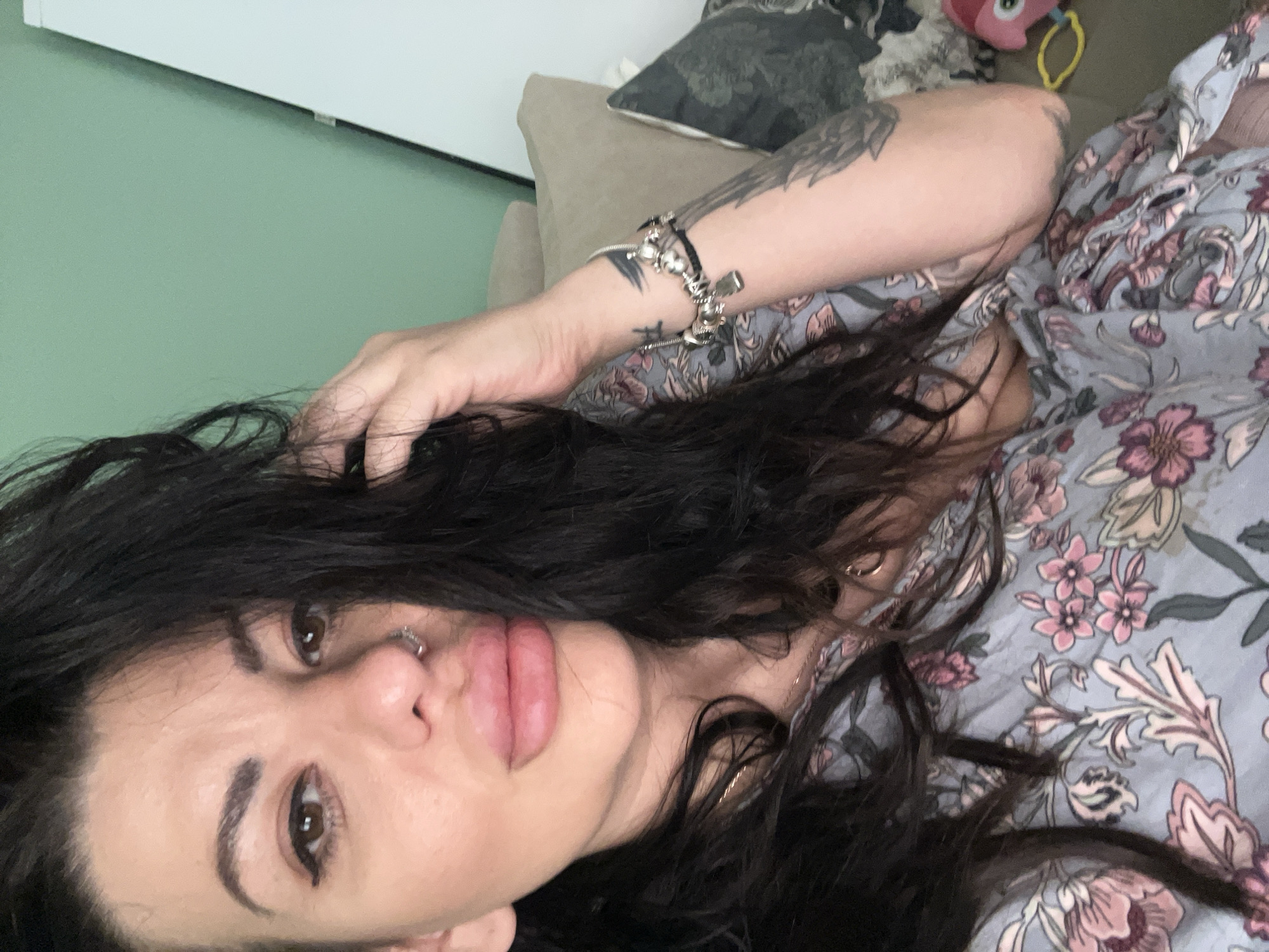 A woman lies on a surface with a floral-patterned fabric. She has dark, wavy hair, brown eyes, and a nose piercing. Her left arm, featuring a tattoo and a charm bracelet, is raised. Behind her are a beige cushion and a patterned pillow. The background includes a light green wall. The location is not possible to determine to be Plovdiv, Bulgaria from the image.