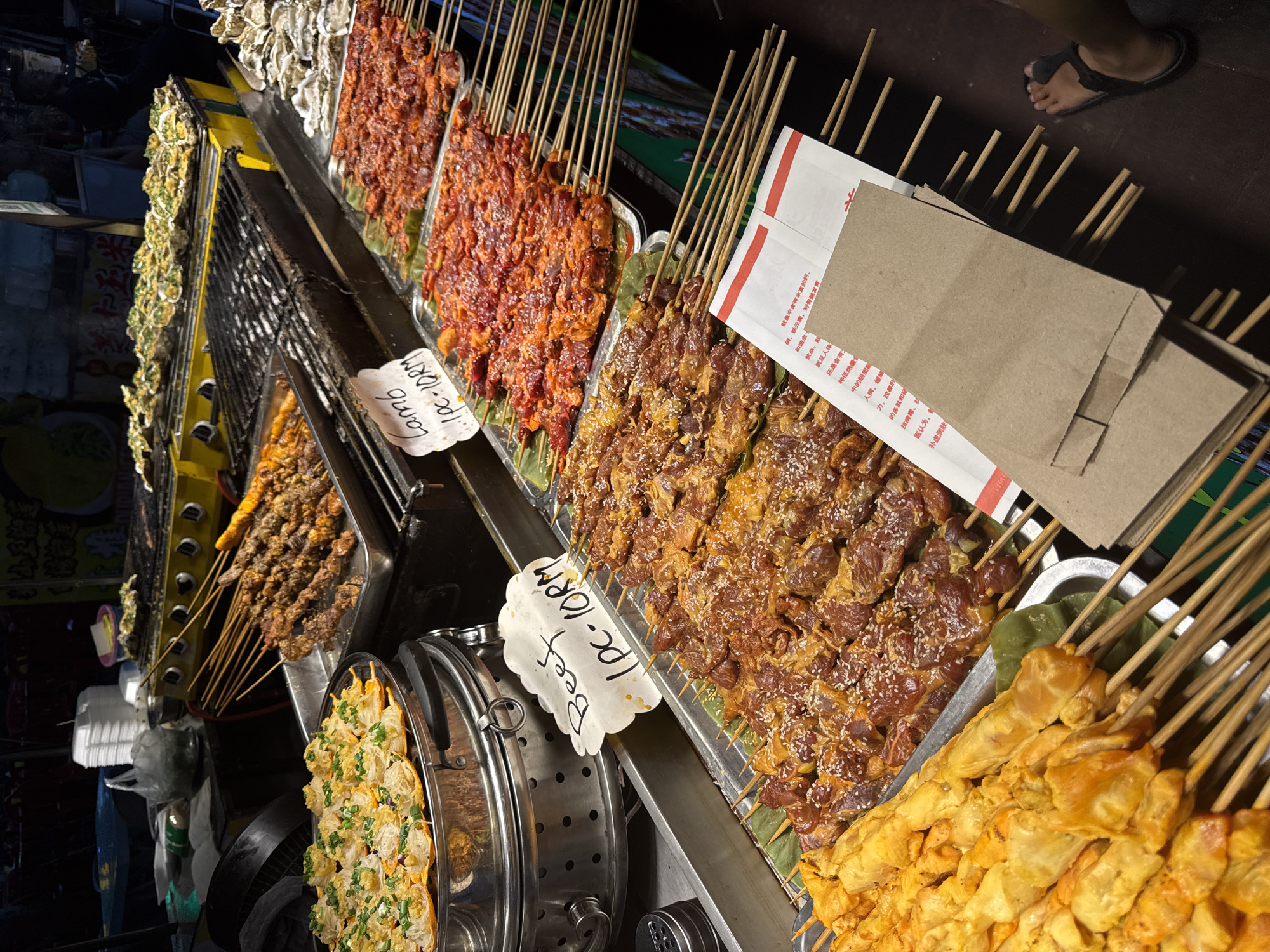 The image captures a vibrant night market food stall, likely in Kuala Lumpur, Malaysia, displaying an array of skewered meats and seafood under artificial lighting. The scene suggests a bustling evening atmosphere.
**Subjects**: The primary subjects are numerous skewers of pre-marinated or raw food items. Rows of lamb skewers, featuring a reddish marinade, are prominently displayed. Adjacent to these are beef skewers, distinguishable by their browner marinade and white sesame seed topping. Further along the stall, there are skewers of what appears to be yellow-colored chicken satay or similar grilled items. On the far left, trays hold what look like grilled oysters or mussels covered in green herbs. A large metal container, possibly a steamer, holds a dish garnished with chopped green onions. Brown paper bags, likely for serving food, are stacked at the back of the stall. A person's foot, wearing a dark sandal, is visible in the upper right corner, indicating human presence.
**Activity or event taking place**: The image depicts a food vendor's stall set up for selling street food. The arrangement of skewers suggests they are ready for grilling or have been freshly prepared for customers. The presence of paper bags indicates items are ready for takeout.
**Notable details**: The setting is clearly a night market due to the dark background and the reliance on artificial lighting to illuminate the food. The food items are colorful and appear well-seasoned. The stalls feature grills and serving equipment. The currency "RM" (Malaysian Ringgit) explicitly ties the location to Malaysia, consistent with Kuala Lumpur.
**Visible text**: Two handwritten white signs provide pricing: "Lamb 1pc - 10RM" and "Beef 1pc - 10RM," indicating that one skewer costs 10 Malaysian Ringgit for both lamb and beef. Additionally, a section of Chinese text is visible, which describes the health benefits of squid/cuttlefish (魷魚), mentioning its rich content of calcium, phosphorus, and iron for bone development, its ability to address anemia, its low-calorie nature, and its benefits for relieving fatigue, improving liver function, providing antiviral properties, and nourishing the skin. This suggests that squid or other seafood might also be offered at the stall, or it's general health information.