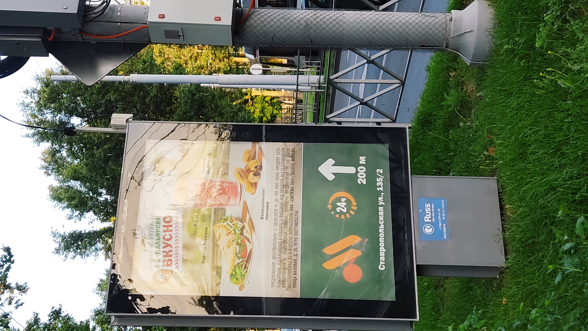 This is a photograph of a billboard advertising food items, likely from a fast-food establishment. The billboard displays an image of a sandwich and what appear to be churros, alongside text in Russian. The text indicates that the offer is valid until May 28, 2026, and that product appearance may differ from the advertisement. It also provides contact information and an address in Moscow for the relevant consumer protection authority. At the bottom of the billboard, there is the logo of a restaurant chain (likely McDonald's, given the M-like symbol and the Russian text "ВКУСНО" meaning "tasty") along with icons indicating 24-hour service and a distance of 200 meters. The address of the specific location is "Ставропольская ул., 135/2" (Stavropolskaya Street, 135/2). This location is in the city of Krasnodar, Russia, as indicated by the street name which is a common street in many Russian cities, and the presence of a McDonald's restaurant at this address. The billboard is mounted on a grey metal pole, which also houses electrical equipment. The scene is set outdoors, with trees and a grassy area in the background. The ground in the foreground is covered with green grass, and there is a metal fence visible to the right. The weather appears to be clear and the lighting suggests it is daytime.