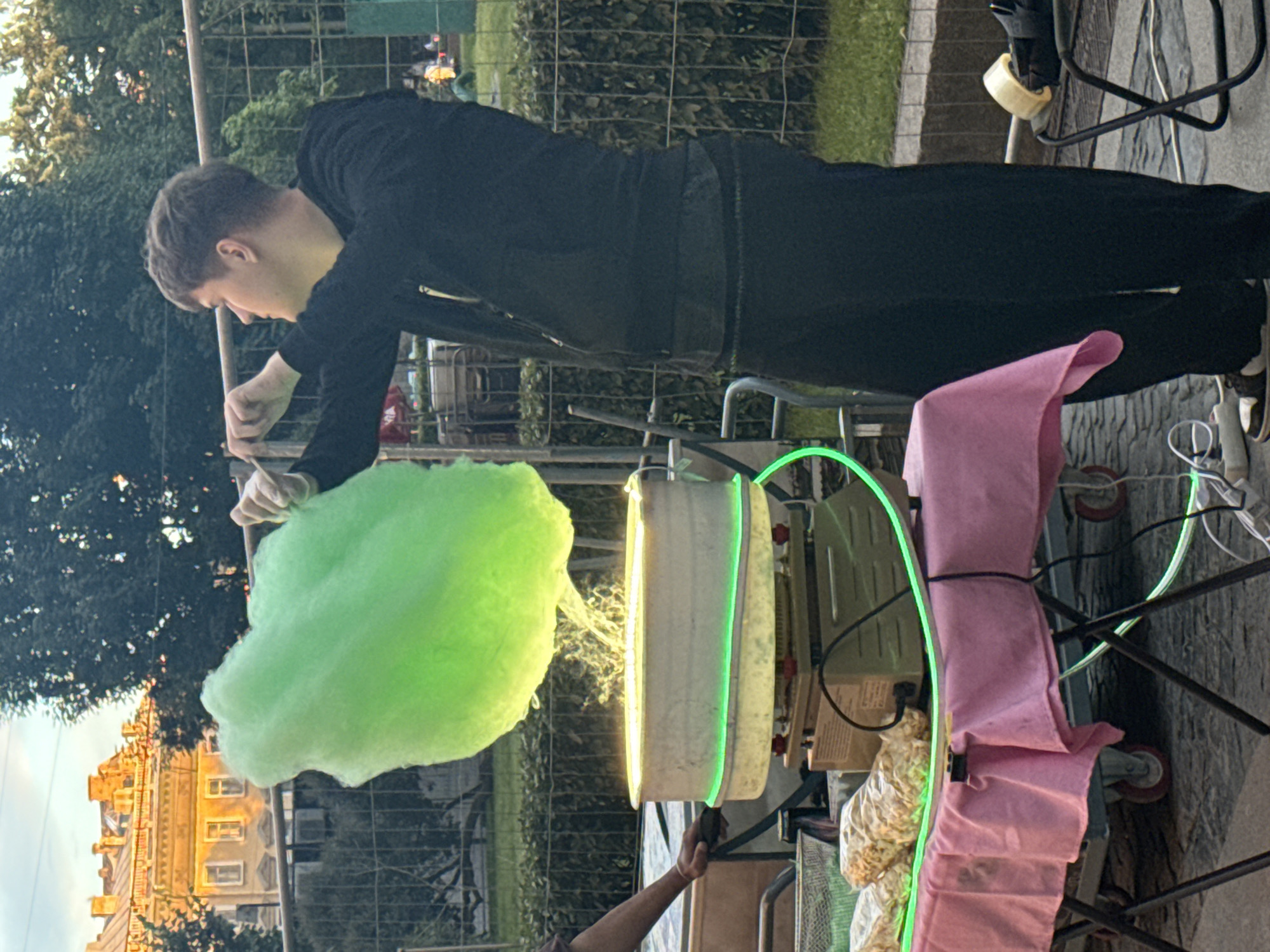 A young man in a black outfit is making green cotton candy from a cart in an outdoor setting. The cart has a bright green neon light around the top of a cylindrical machine. Pink fabric is draped over part of the cart. In the background, there is a building with many windows, suggesting an urban environment. Trees are also visible, indicating the presence of nature. The scene appears to be taking place during the day, possibly late afternoon or early evening, given the lighting. The man is focused on the task of creating the cotton candy, and the overall atmosphere seems casual and perhaps festive. The image is oriented vertically, and the ground is paved with stone. No visible text is present in the image.