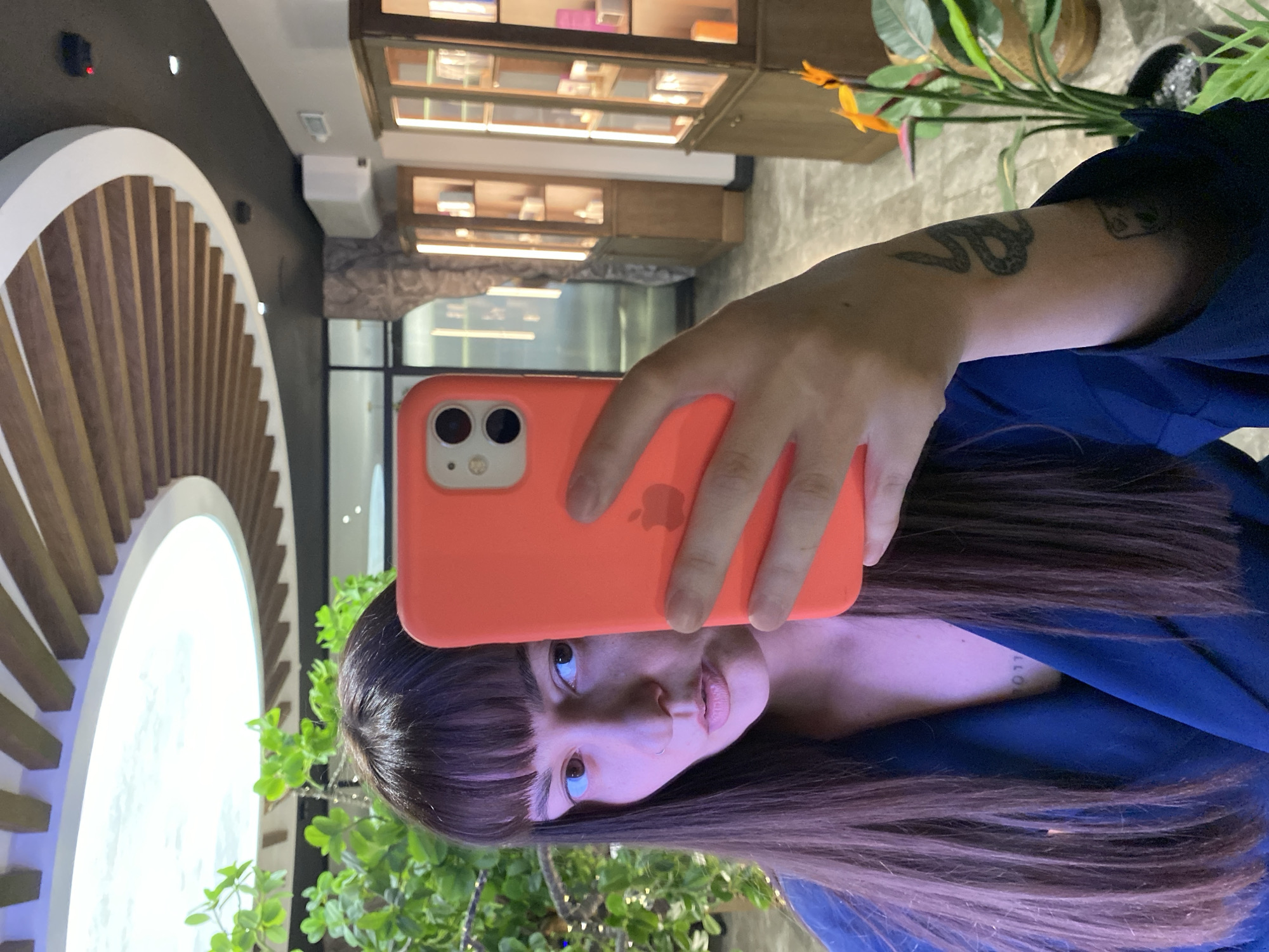 An inverted selfie captures a young person in an indoor setting, likely in Votkinsk, Russia. The individual, with long dark hair and bangs, looks directly at their phone, which they are holding up to take the photo. They wear a dark blue top or jacket.
The smartphone is a bright coral/orange color, with the Apple logo visible on its back and a dual-lens camera system. Notable personal details include a snake tattoo on the person's right forearm (viewer's left) and a partially visible tattoo of text or numbers, possibly "2017," on their left shoulder/upper arm.
The background features modern interior design elements. To the left, a large, illuminated circular architectural feature with radiating wooden slats creates a striking visual. In the background, there are wooden display cases or shelves containing various items. Green plants with orange-yellow flowers are visible in the foreground and mid-ground, adding greenery to the space. The overall lighting suggests an indoor setting, possibly a lounge, cafe, or retail area, with artificial light illuminating the scene. The person's expression is neutral as they engage in the act of taking a selfie.
