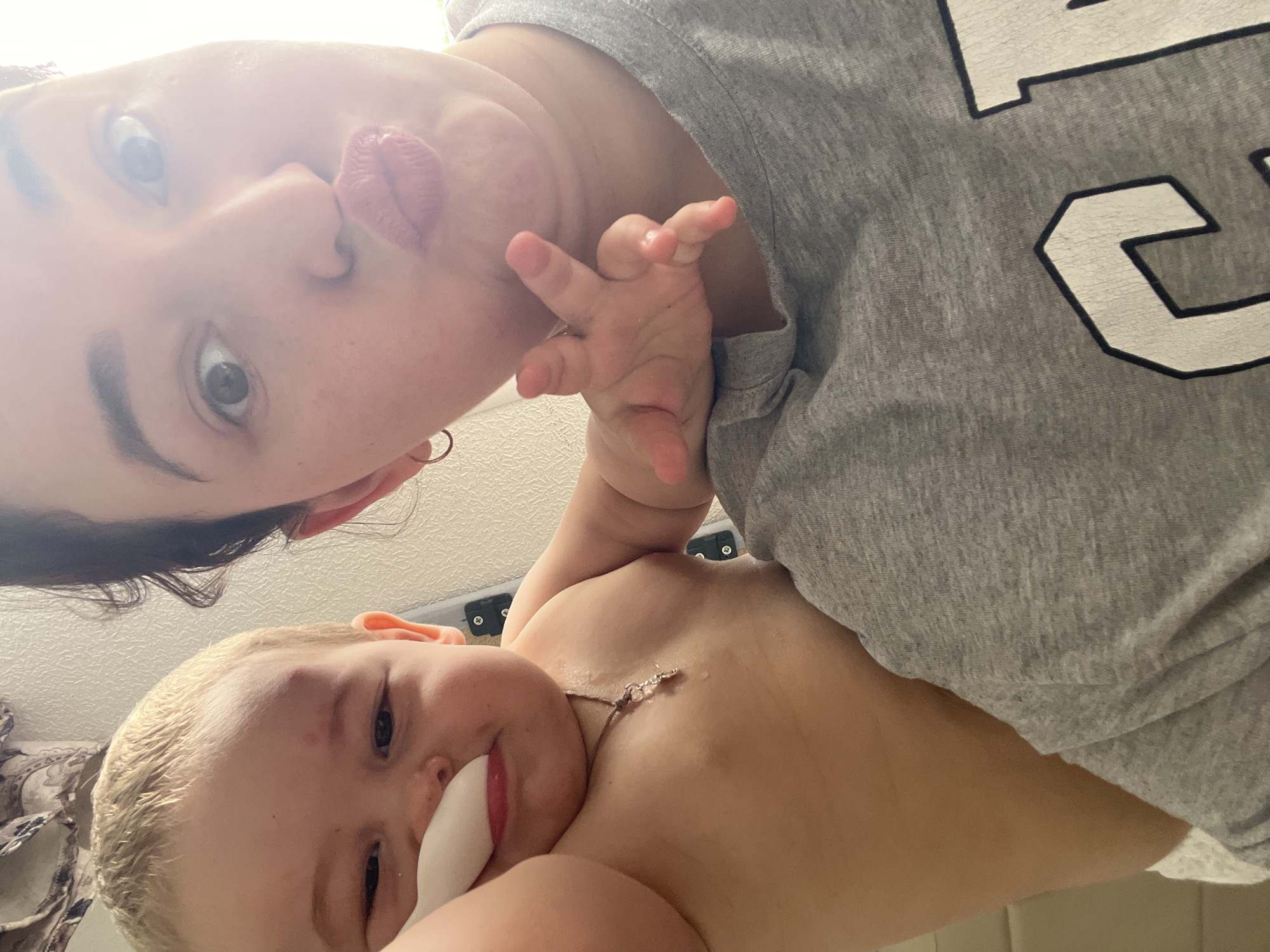 This is a selfie taken indoors, likely in a domestic setting, featuring a woman and a baby. The woman is in the foreground, making a kissing face towards the camera. She has dark hair and is wearing a grey t-shirt with a logo partially visible. A baby is positioned behind her, leaning forward and touching the woman's shoulder. The baby has blonde hair and is wearing a necklace. The background appears to be a plain wall. There is no indication of the location being in Chelyabinsk, Russia.