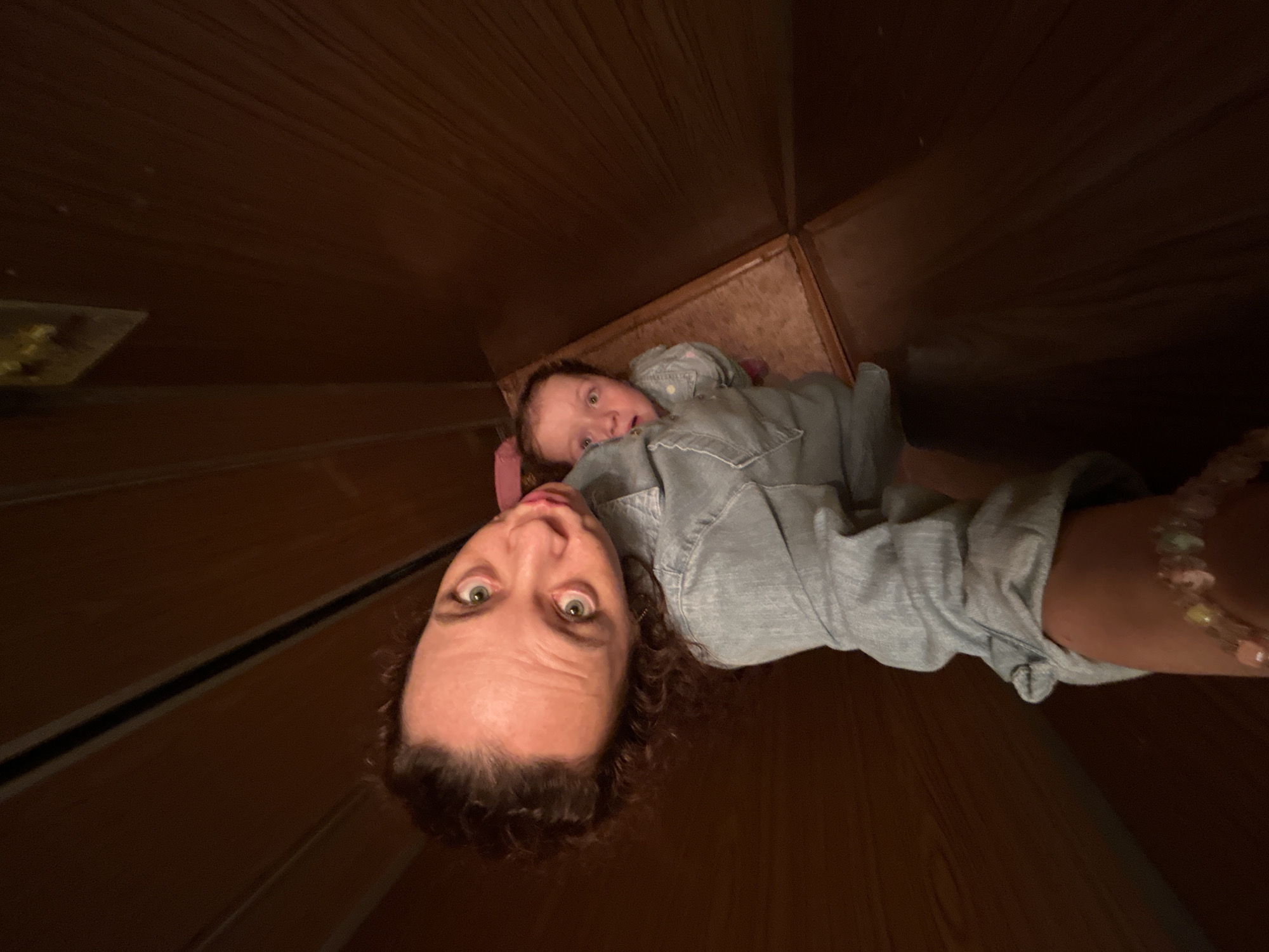 This media file is a selfie taken in a dimly lit, wooden interior. The main subjects are a woman and a baby. The woman, positioned in the foreground and upside down, has curly brown hair and wide green eyes with furrowed brows, conveying surprise or alarm. She is wearing a light blue denim shirt and a beaded bracelet on her right wrist. The baby, lying behind her and also upside down, has light hair and is looking upwards. The baby is also wearing a light blue denim shirt.
The setting appears to be a confined space with wooden paneling on the walls and floor, suggesting it might be a closet, an attic, or a similar narrow area. The wooden elements have a warm, brown tone. There are some faint horizontal lines or seams visible on the wooden surfaces.
The activity captured is a selfie, likely taken by the woman. The unusual perspective and the expressions on both subjects' faces suggest a playful or perhaps a slightly mischievous moment, though the woman's expression could also indicate genuine surprise at the camera's positioning. The time of day is not clearly indicated, but the lighting suggests an indoor setting, possibly at night or in a room with limited natural light. There is no visible text in the image. The location context is Chaikovsky, Russia, but there are no specific visual cues in the image to confirm this.