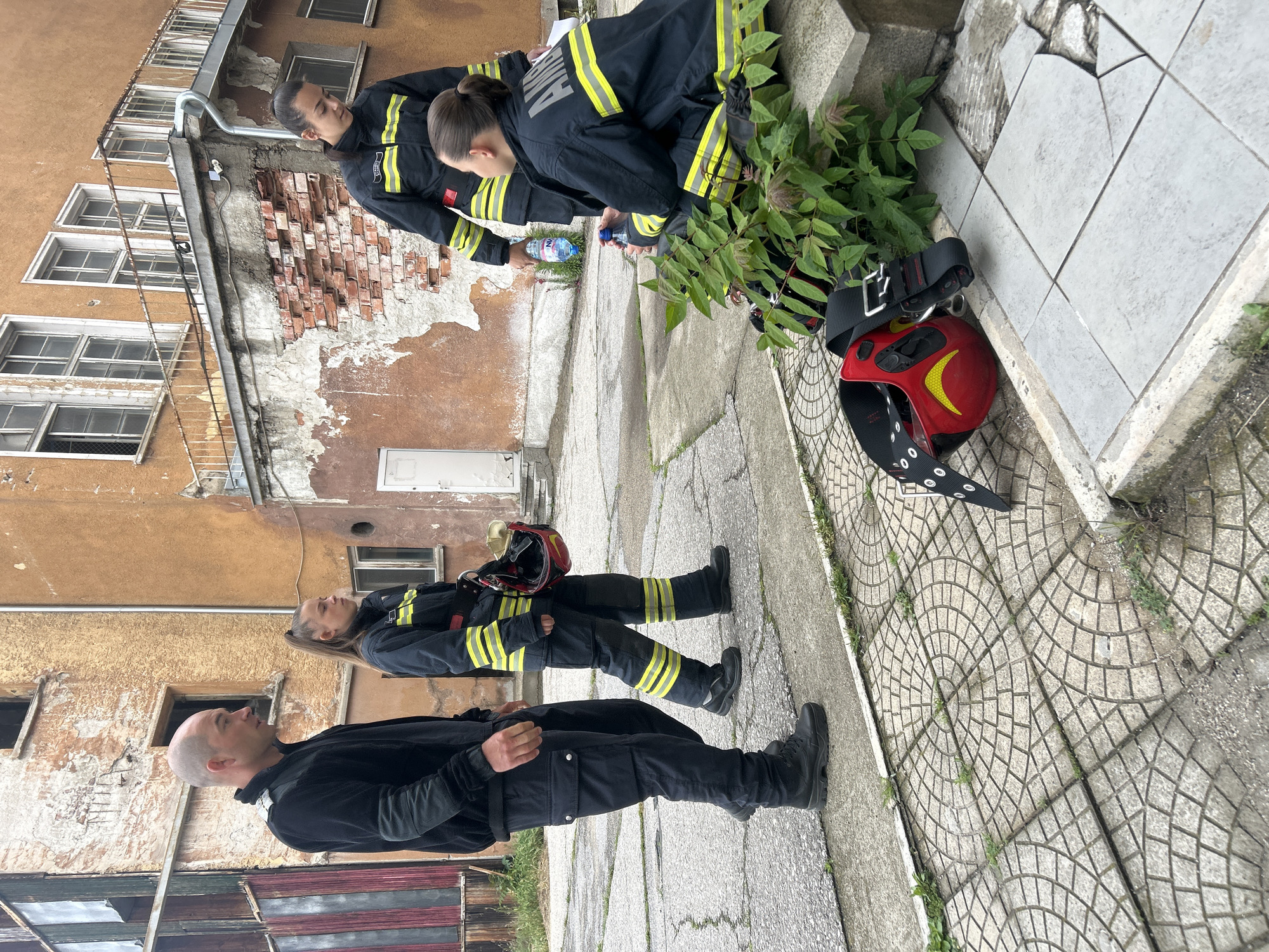 The image depicts four emergency responders in what appears to be an urban setting, likely Sofia, Bulgaria, under daytime, possibly overcast, conditions.
Three individuals are dressed in dark-colored uniforms with reflective yellow-green stripes, characteristic of emergency services. One female responder, seen from the back, has "AMBULANCE" clearly visible on her uniform's shoulders, indicating medical emergency personnel. She is kneeling near a patch of greenery, holding a "NUI" branded water bottle, possibly hydrating or sharing it. Another responder in similar attire, a female with her hair in a bun, stands nearby, observing. A bald male in a plain black uniform stands in the foreground, looking upwards and to the right, his hands clasped.
Two red emergency helmets are present, one with a yellow crescent logo resting on the ground next to a black harness, and another being held by a responder.
The background features an old, multi-story building with a faded orange-brown facade. The building shows signs of disrepair, including exposed brickwork where plaster has peeled off, numerous small, rectangular windows, and a metal drainpipe. The ground is paved with concrete, some sections featuring a decorative circular pattern, with weeds growing through cracks and along concrete steps. The scene suggests a break or an assessment during an operation in a somewhat aged urban environment.