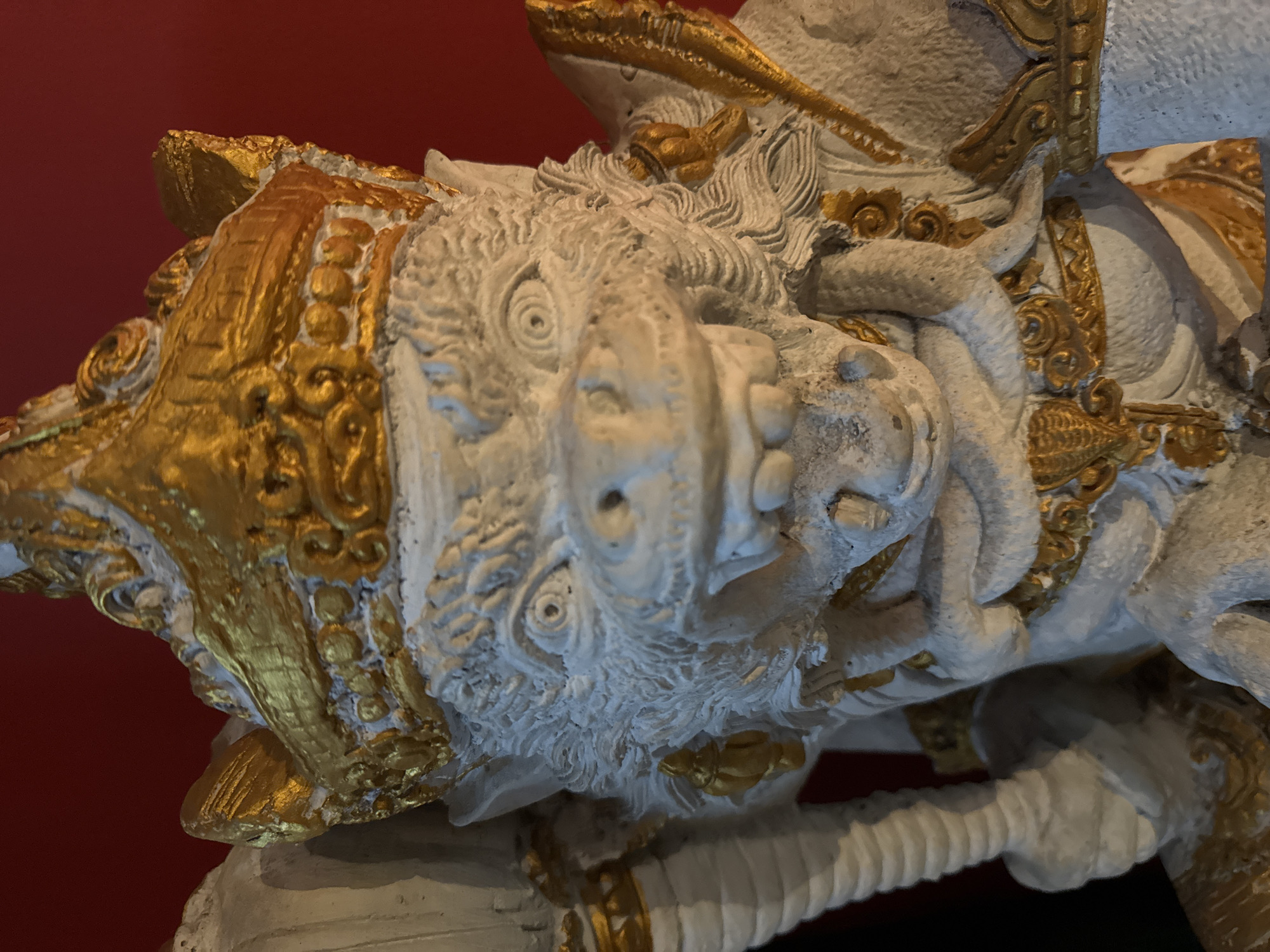 This is a close-up, eye-level photograph of a white and gold ornate sculpture. The sculpture depicts a creature with a wrinkled snout, bulging eyes with concentric circles for pupils, and a mane of textured hair. It is adorned with elaborate gold detailing, including decorative bands around its head and body, and what appear to be stylized floral or leaf motifs. The creature's form is somewhat rough and textured, suggesting it might be made of plaster or a similar material. One of its forelimbs, rendered in a ribbed, tubular style, is visible in the lower portion of the frame, also with gold accents. The background is a plain, deep red, which contrasts with the lighter tones of the sculpture. Given the location context of Kuta, Indonesia, this sculpture likely represents a mythical creature, possibly a guardian or decorative element found in temples, palaces, or traditional Balinese architecture. The lighting appears to be artificial, casting subtle shadows and highlighting the textures and gold embellishments. No text is visible in the image.