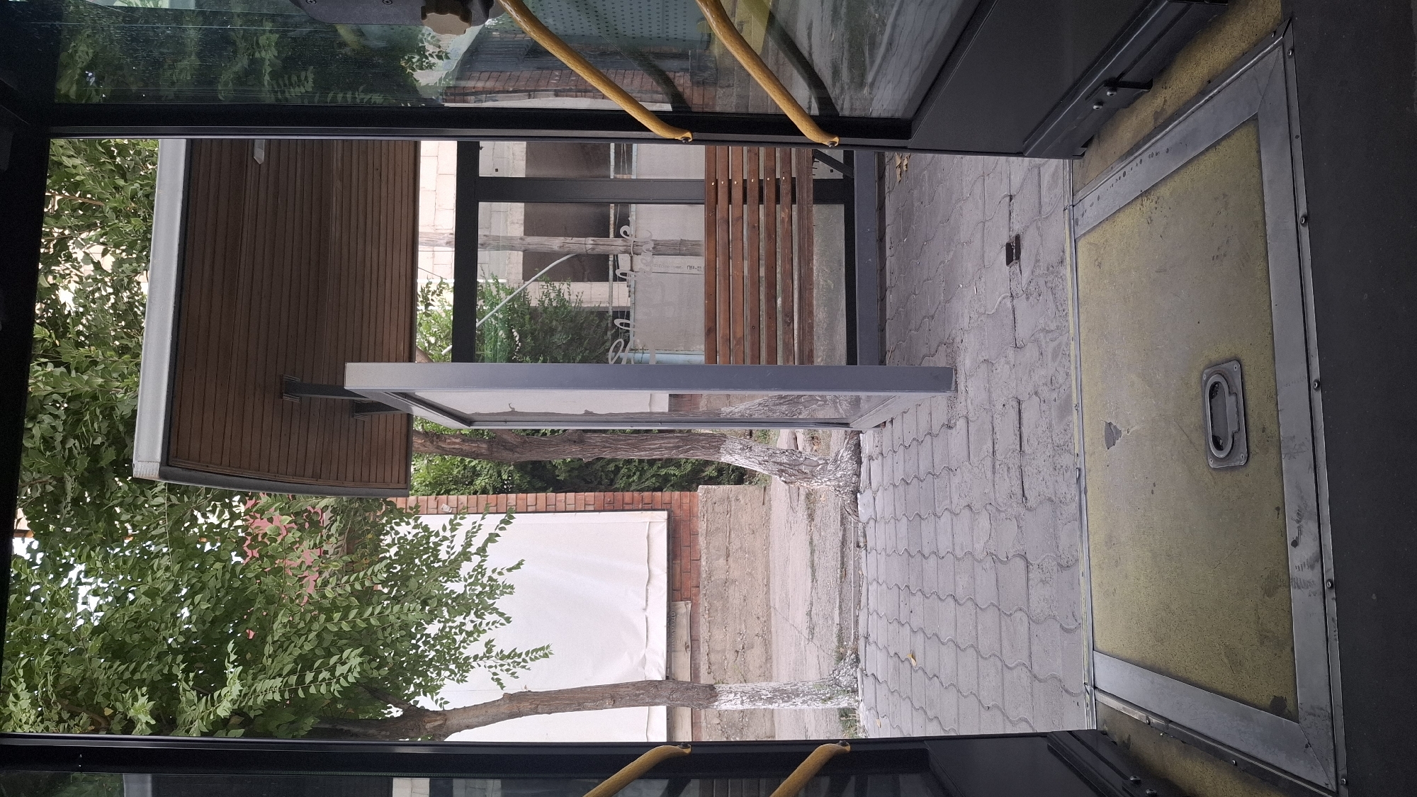 A view from inside an open bus door reveals a bus stop shelter. The bus floor, with a yellow mat and a metal handle, is in the foreground. The bus stop shelter has a wooden slatted roof and a glass enclosure with a wooden bench. Two trees with whitewashed trunks are visible outside the shelter. The ground is paved with interlocking stones. In the background, a brick building with a white tarp is seen. The location is Shymkent, Kazakhstan.