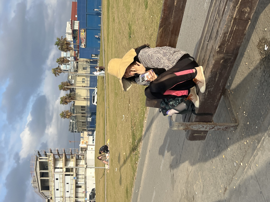 A woman in a wide-brimmed hat, patterned sweater, and black leggings with a pink stripe sits cross-legged on a wooden bench, using her smartphone. A backpack is visible beneath her. The background shows a grassy area, buildings, palm trees, and a cloudy sky. The bench is on a paved path, with a partially constructed building in the distance. The image is rotated.