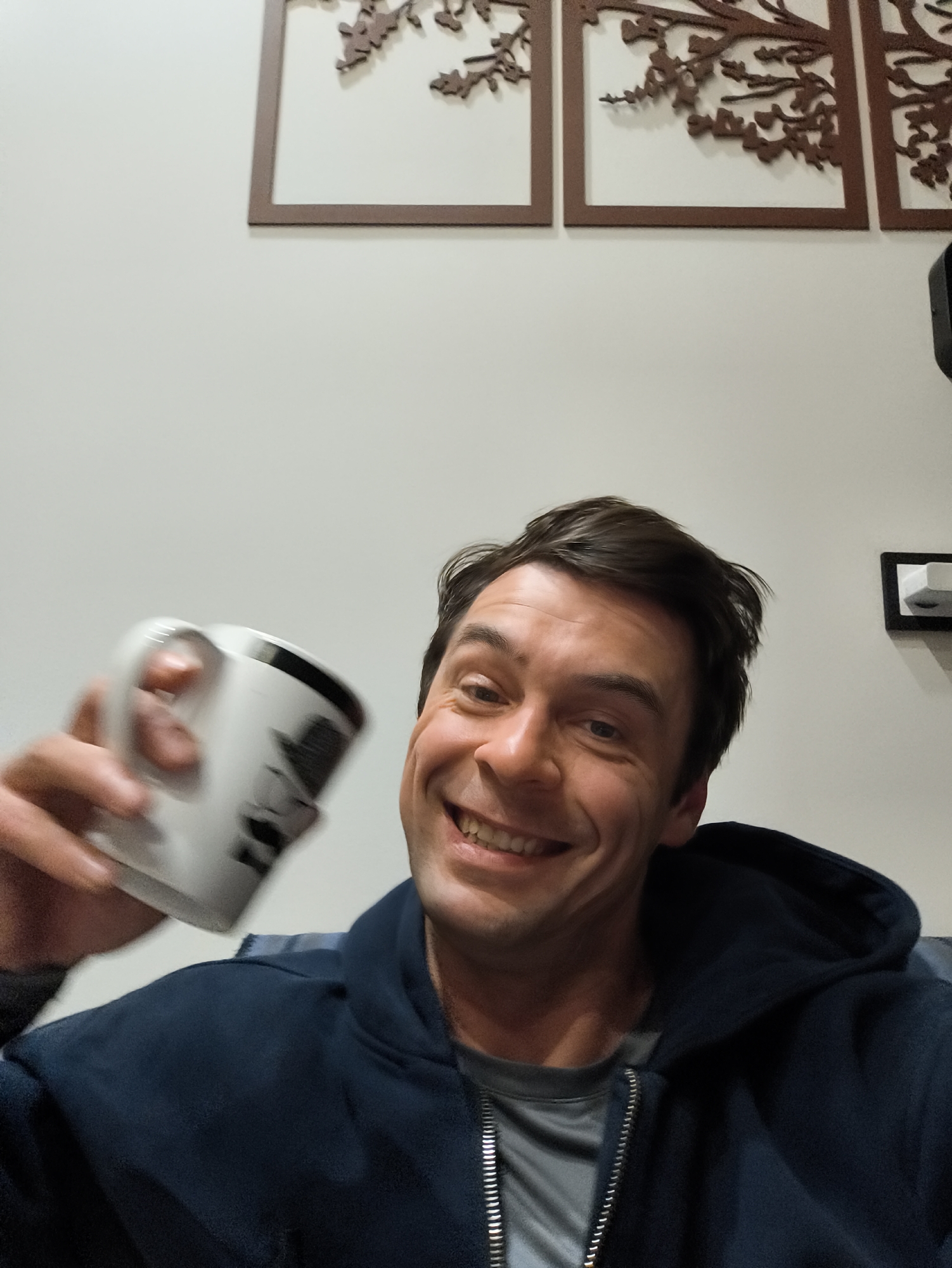 A selfie shows a smiling man in a blue hoodie holding a white mug. Behind him is a white wall with three framed tree branch designs and a black object with a white piece attached to the wall. The location is Unknown, Unknown.