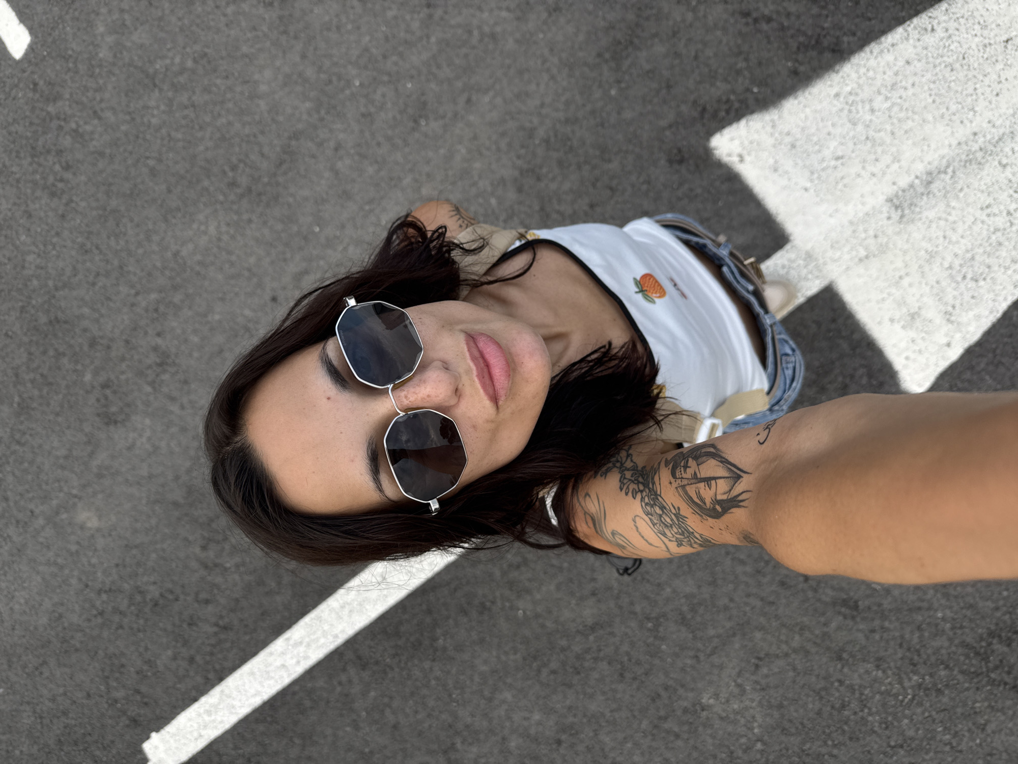 A high-angle selfie captures a young adult female on an asphalt surface in Pleven, Bulgaria. She has dark hair, freckles, and wears octagonal silver-framed sunglasses with dark lenses reflecting a cloudy sky. Her lips are slightly parted. She is dressed in a white tank top featuring an orange fruit emblem and partially visible blue denim jeans with a brown belt. Her right arm is visible, heavily tattooed with intricate designs, including a split-face motif on the inner bicep and what appears to be Arabic script near the elbow. The subject is looking upwards towards the camera. The background consists of dark grey asphalt with prominent white painted road or parking lines, one running diagonally across the upper left and another forming a perpendicular angle in the lower right.
