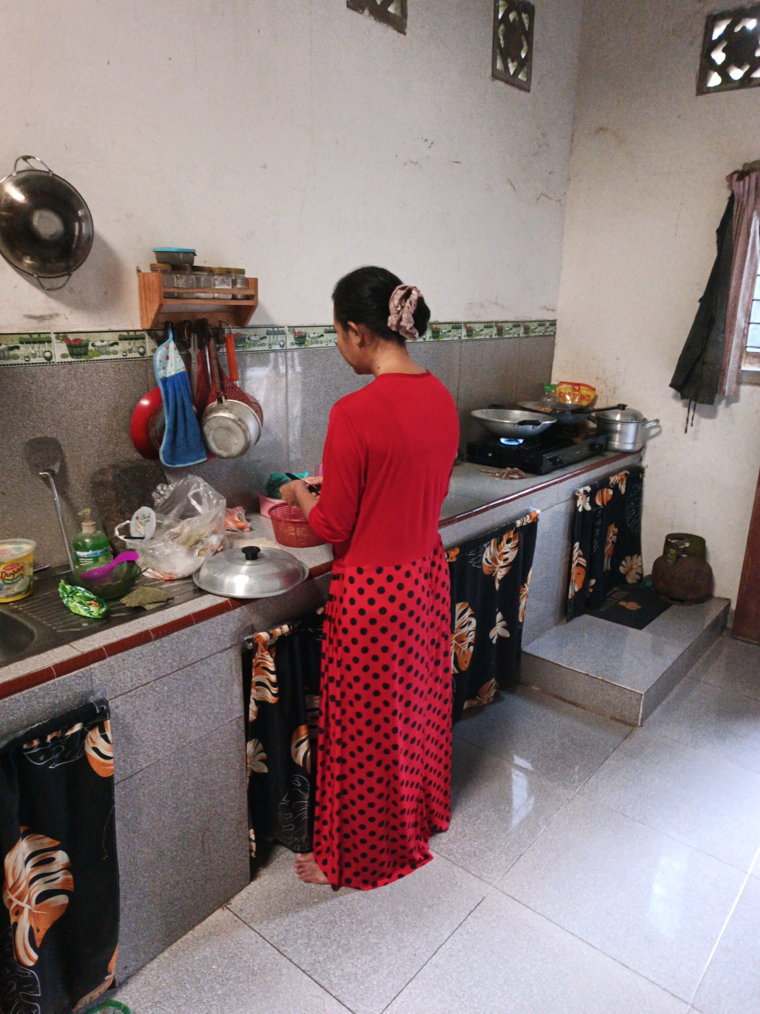 An Indonesian woman with dark hair tied in a bun is standing in a kitchen, facing away from the camera. She is wearing a red long-sleeved top and a long red skirt with black polka dots. She is holding a red bowl and appears to be preparing food. The kitchen has white walls and grey tiled countertops. Various kitchen utensils and appliances are visible, including pots, pans, a stove, a sink, and a dish rack. There are also decorative elements such as patterned curtains and a small shelf with spice jars. The floor is tiled in a light grey color. The overall impression is of a functional, lived-in kitchen.