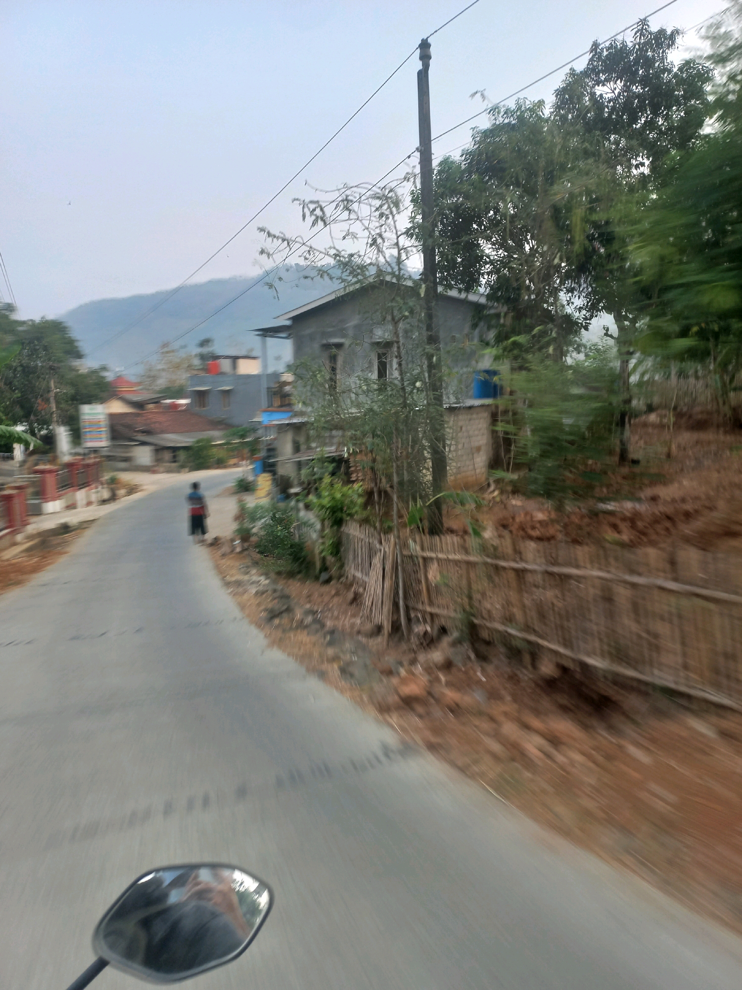A lone figure walks down a paved road in a rural area of Sukoharjo, Indonesia. The person, wearing a dark shirt with horizontal stripes and dark shorts, is in the middle of the road, facing away from the camera. The road curves to the left, flanked by a dirt embankment on the right and a sidewalk on the left. On the right side of the road, a rustic bamboo fence runs along the embankment, with trees and vegetation behind it. Further back, a single-story house with a grey concrete exterior and a blue water tank on the roof is visible. The background shows a hazy sky, distant hills, and more houses, indicating a small village or settlement. The image appears to be taken from a moving vehicle, suggested by the motion blur on the road and surrounding environment, and the presence of a motorcycle's rearview mirror in the bottom left corner, reflecting the photographer. The overall atmosphere is one of a quiet, early morning or late afternoon in a semi-urbanized rural setting.
