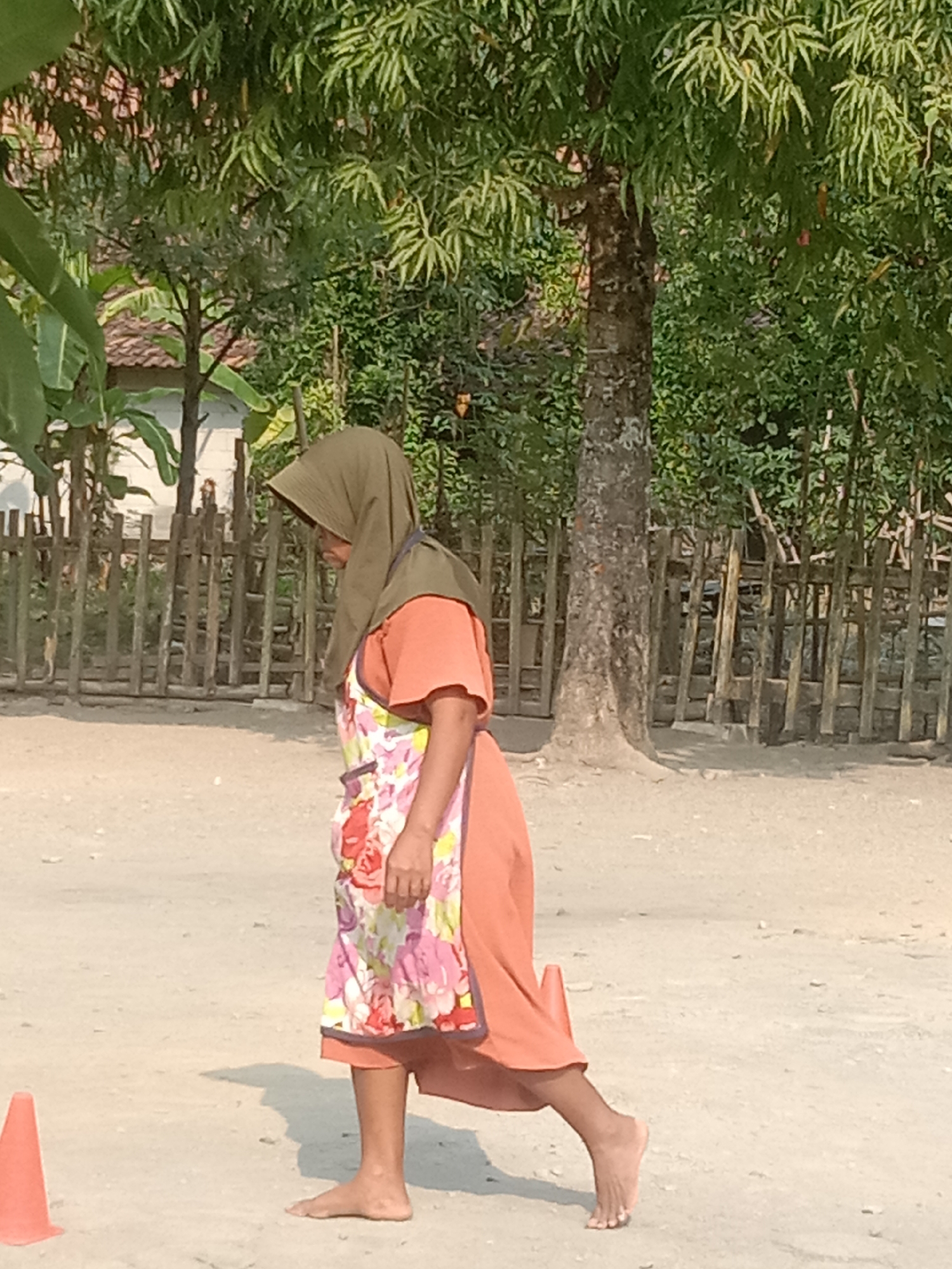 An elderly woman in a headscarf and an orange dress, wearing a floral apron, walks barefoot on a dirt road in Tanjungsari, Indonesia. She is in profile, facing left, with her right leg forward in a walking stride. To her left, a small orange cone is visible on the ground. Behind her, a wooden fence encloses a yard with trees and vegetation. The scene appears to be outdoors in a rural or semi-rural setting.