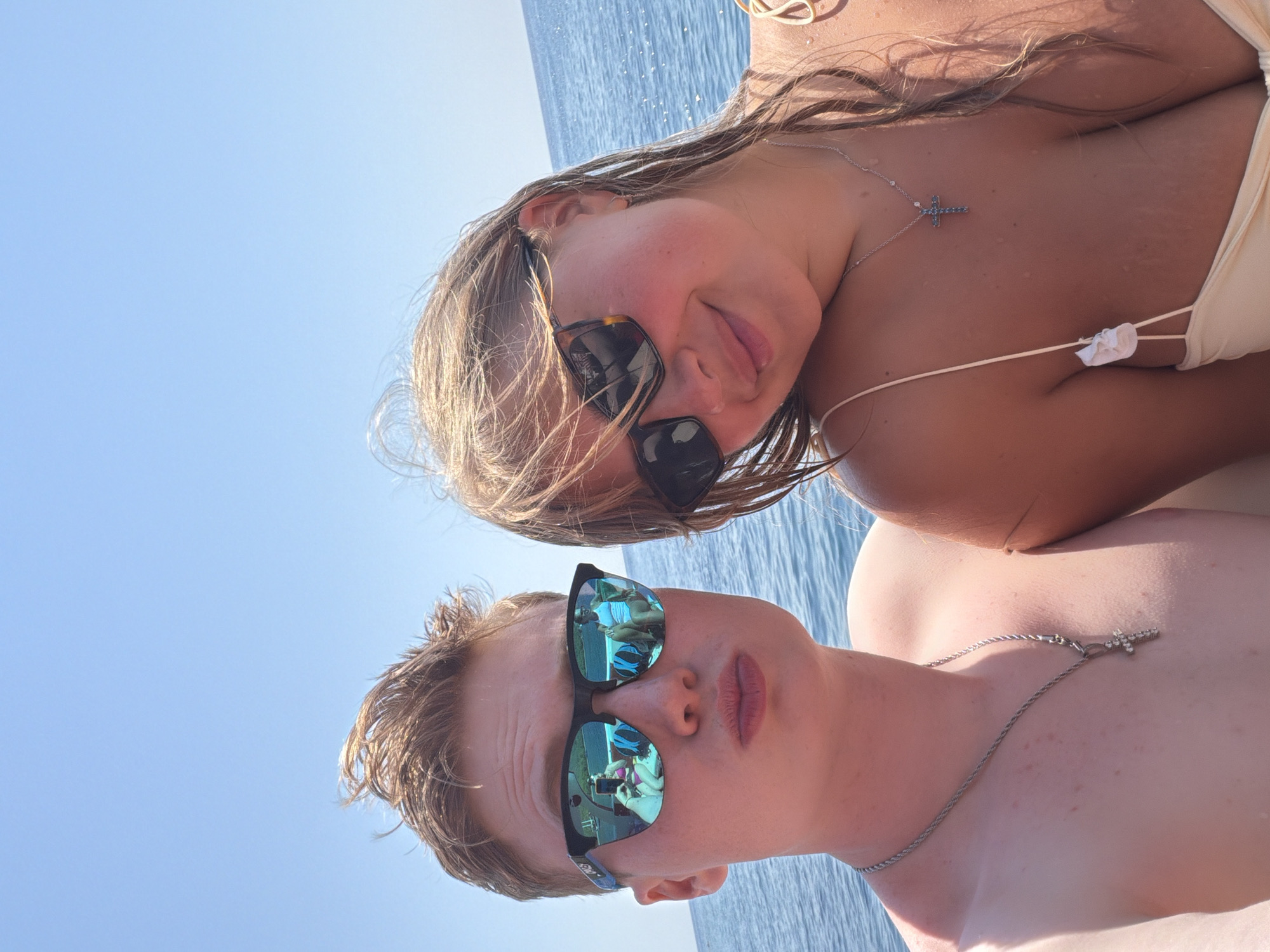 A young Caucasian couple is on a boat trip in the Mediterranean Sea, with the coast of Kemer, Turkey visible in the background. The man, on the left, is wearing sunglasses and a silver chain with a cross pendant, and is making a duck face. The woman, on the right, is also wearing sunglasses and a necklace with a cross pendant, and is smiling at the camera. They are both wearing swimwear. The sun is shining brightly, and the water is calm.