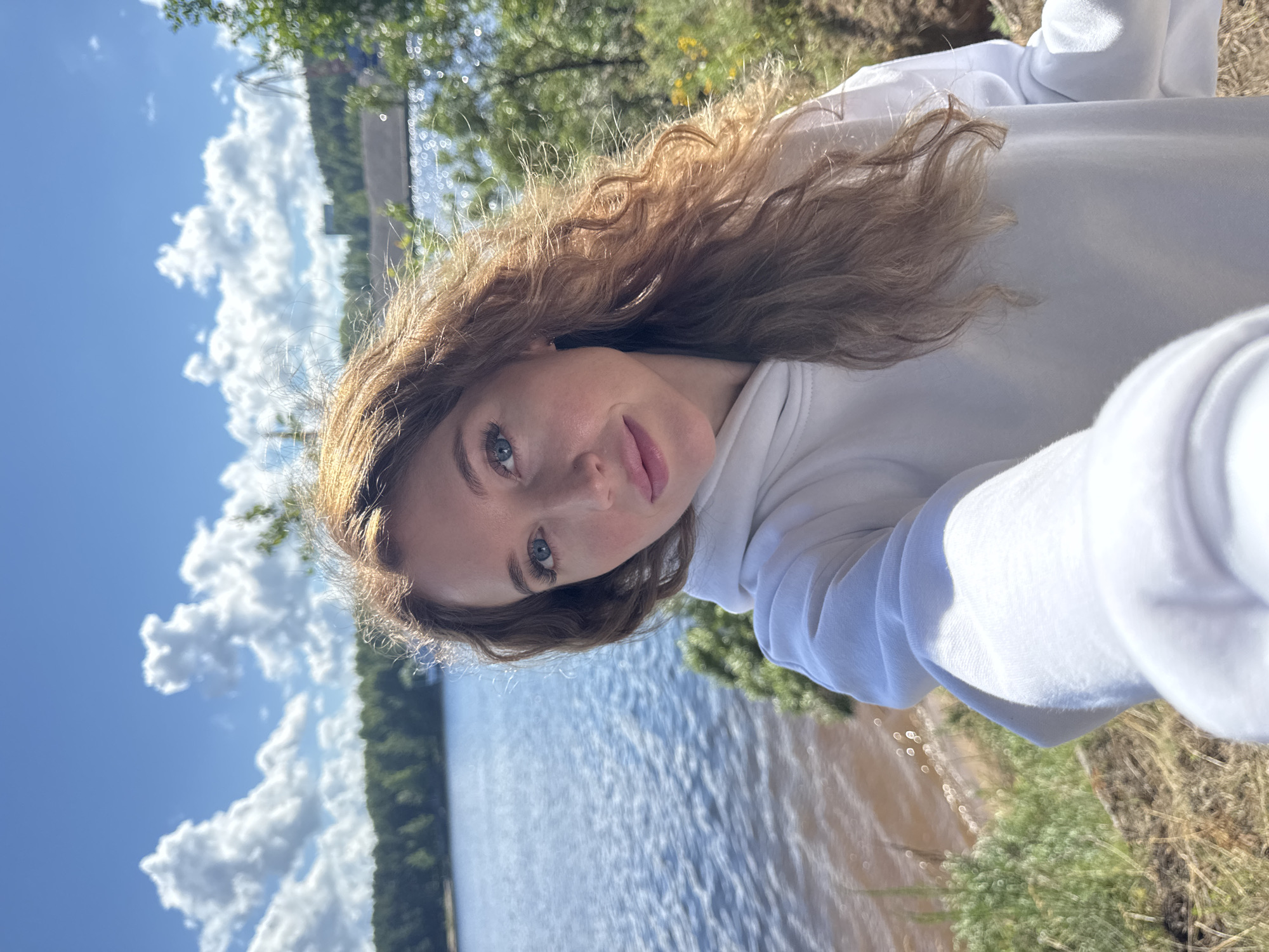 A woman with long, wavy auburn hair takes a selfie outdoors on a sunny day. She is wearing a white hoodie and looking directly at the camera with blue eyes and light pink lips. The background shows a body of water, likely the Kama River, with a forested shoreline and a bright blue sky with fluffy white clouds. In the distance, industrial structures and cranes are visible, suggesting the presence of port or industrial facilities typical of Chaikovsky, Russia. The foreground consists of dry grass and some green bushes, indicating a natural, slightly rugged embankment.