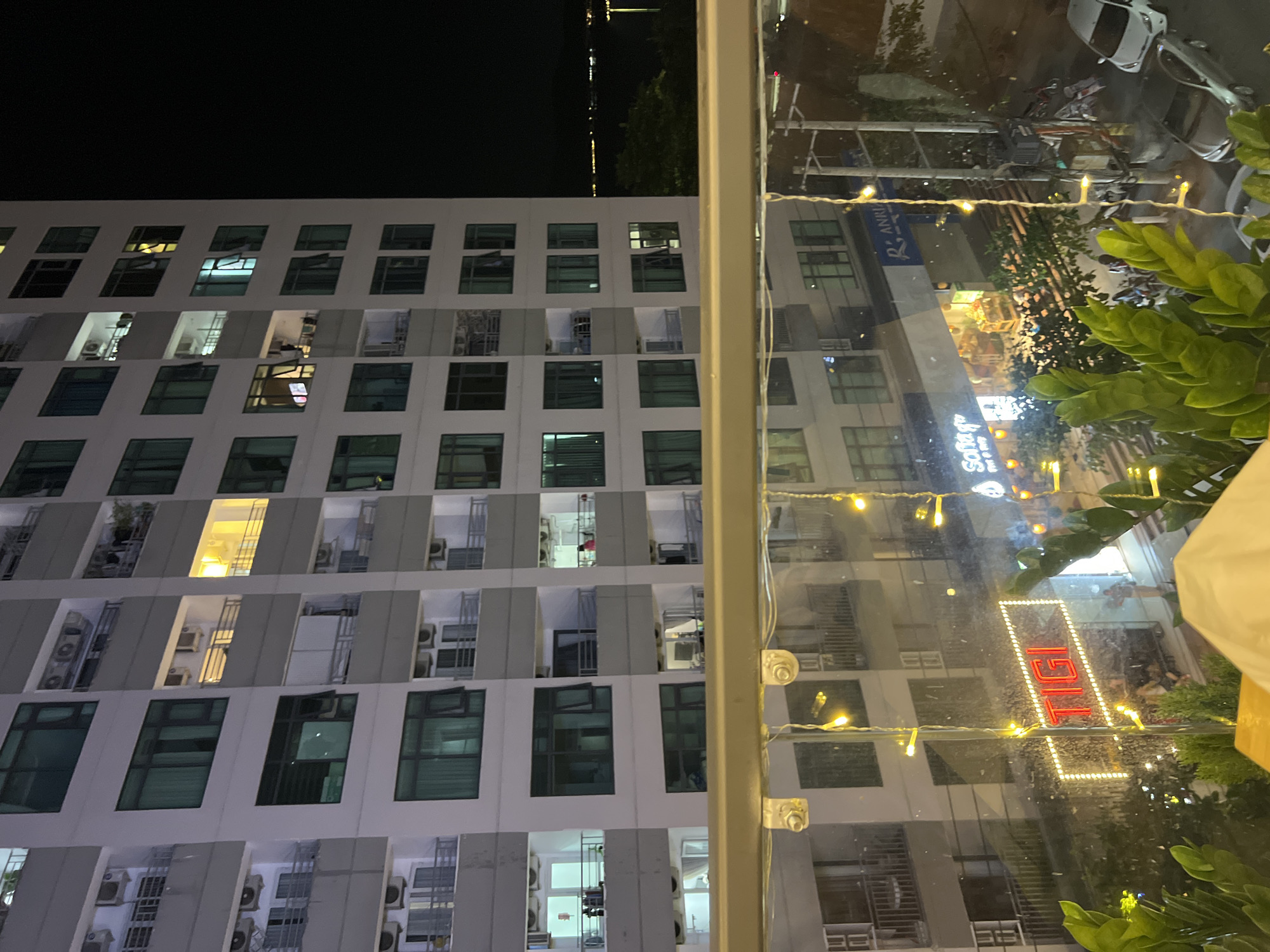 The image presents a nighttime urban landscape, likely in Bắc Nha Trang, Vietnam, captured from a building or balcony viewpoint. The photograph is oriented 90 degrees clockwise.
On the left, a tall, modern high-rise building dominates, characterized by its light-colored facade and a repetitive grid of windows. Many windows are illuminated, indicating active rooms and revealing interior lights and, in some cases, air conditioning units on small balconies. The sky above the building is dark, with distant, faint lights suggesting other structures or the coastline.
The right side of the image is filled with reflections on a glass surface, possibly a window or balcony railing. Decorative string lights are strung across this foreground element, and vibrant green foliage is visible at the bottom right. The reflections reveal a bustling street scene below, featuring illuminated business signs, utility poles with wires, a white car, and parked motorbikes, all pointing to urban activity.
Visible text includes a prominent neon sign reflecting the word "TIGI" in red and white. Other reflected text, though less distinct, includes "B. RANNA" (partially visible) and faintly, "Soho Cafe". The overall scene captures the vibrant atmosphere of a city after dark.
