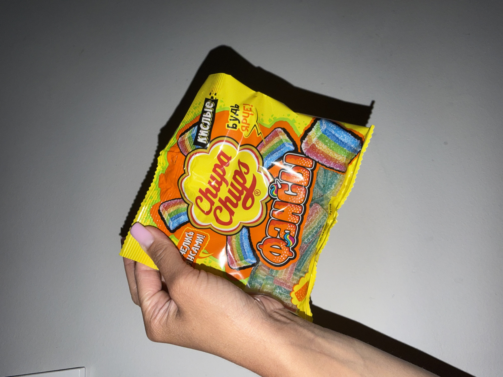 A hand with light skin and a pink-painted fingernail holds a bag of Chupa Chups sour strips. The bag is yellow and orange with a cartoon rainbow motif and Russian text. The text "Chupa Chups" is prominently displayed on the bag. The background is a plain gray wall, creating a studio-like effect. The image is lit from the front, casting a soft shadow to the right of the bag. There are no identifiable landmarks or elements in the image to suggest a location in Sirius, Russia.