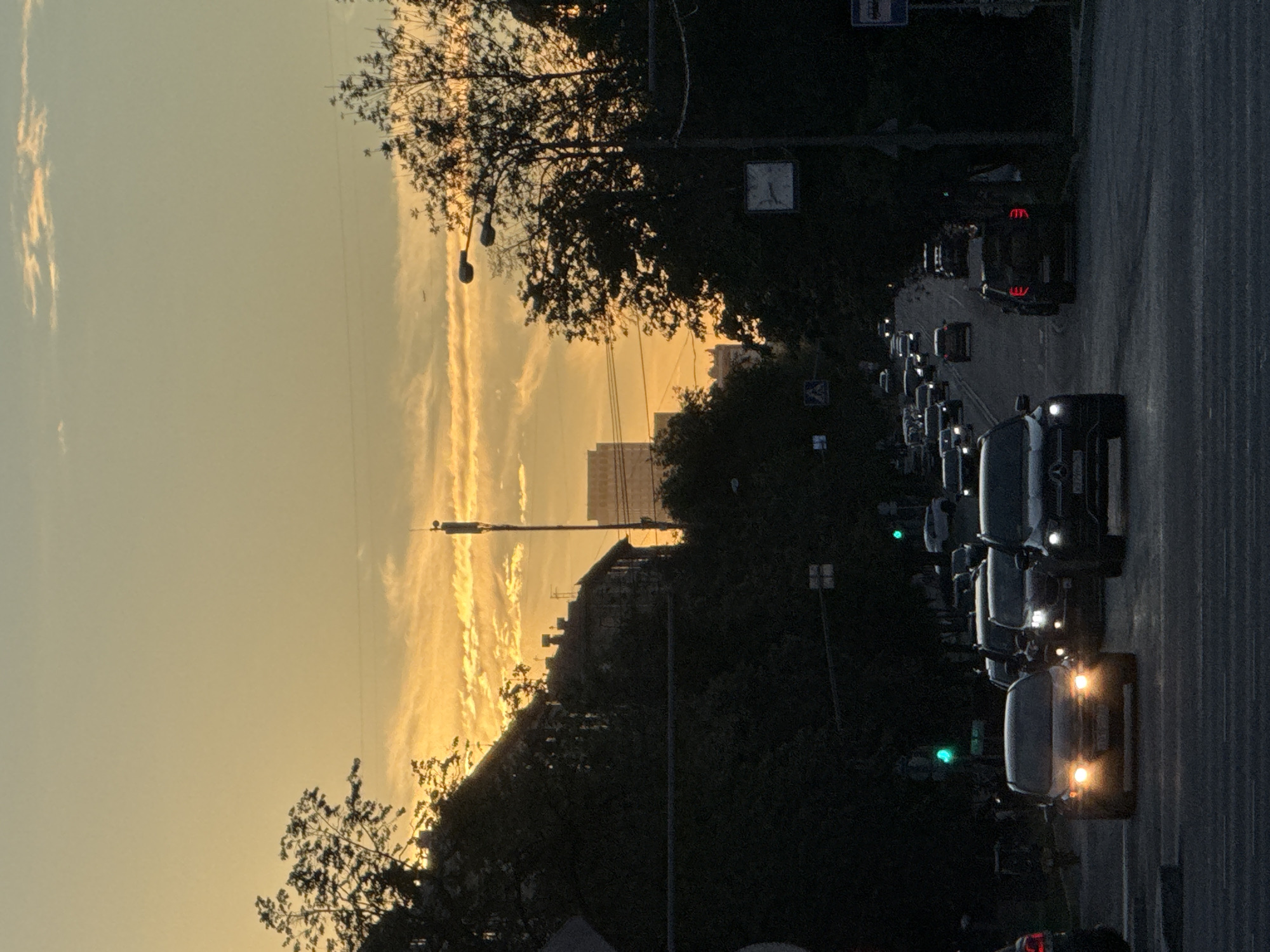 The image depicts a street scene at dusk, with a clear sky filled with wispy, golden clouds. The sun is setting, casting a warm, ethereal glow over the landscape. A line of cars is driving down a paved road, their headlights and taillights illuminated. Trees line the sides of the road, their branches silhouetted against the sky. In the distance, buildings and streetlights can be seen. One of the cars in the foreground appears to be a dark-colored sedan, possibly a Mercedes-Benz, based on the visible branding. The traffic lights are visible, with one showing a green signal. The overall mood of the image is peaceful and serene, evoking a sense of transition from day to night. There is no visible text in the image.