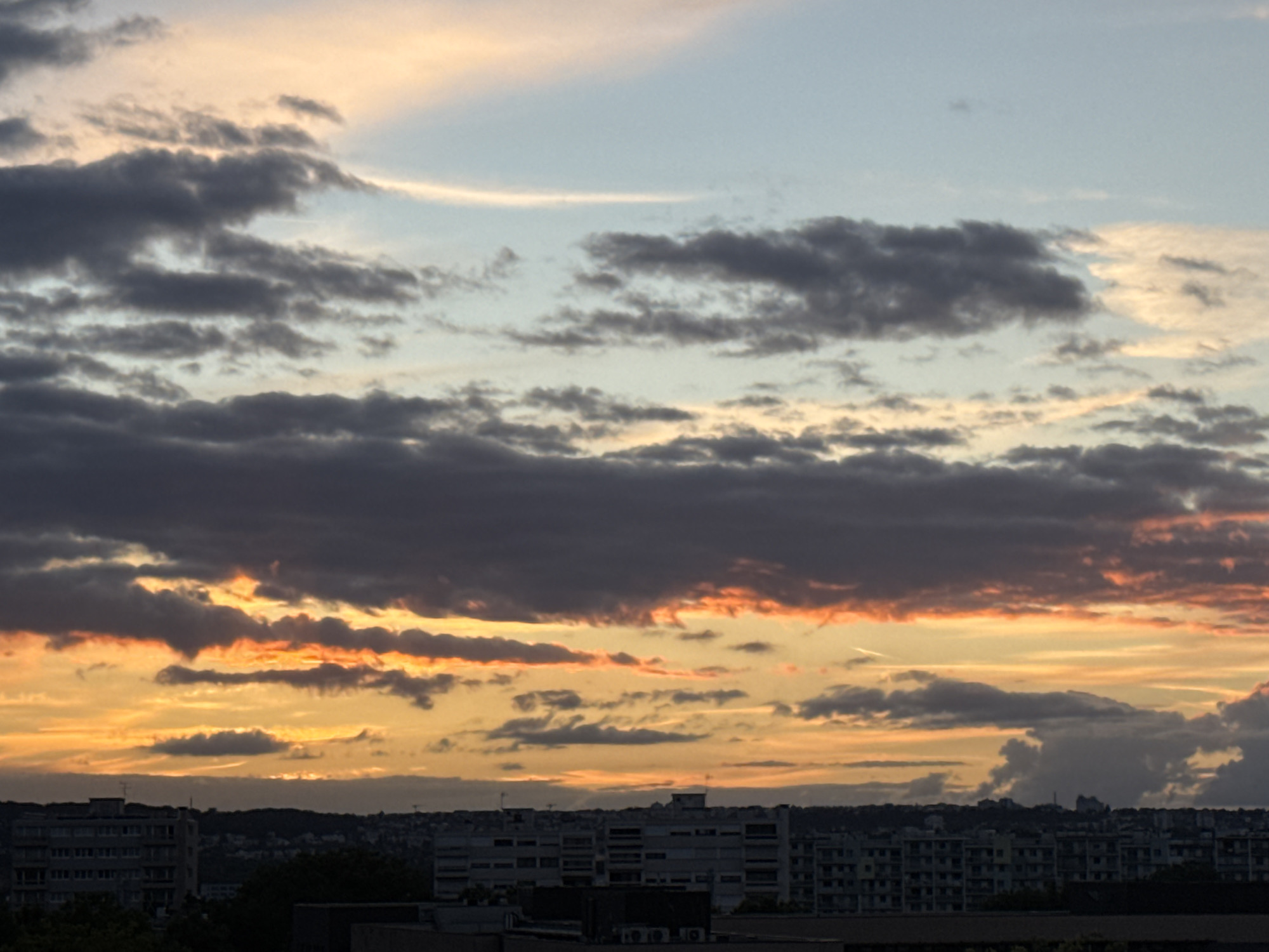 This image depicts a sunset over the city of Fresnes, France. The sky is filled with a variety of clouds, ranging from dark gray to lighter hues tinged with orange and yellow from the setting sun. Some clouds appear wispy, while others are more dense and layered. Below the sky, a cityscape is visible, characterized by rows of apartment buildings. The architecture suggests a modern urban environment. The overall impression is one of a calm, late-evening scene in a suburban area of France. There are no visible people or specific actions taking place. The focus is on the atmospheric conditions and the urban landscape.