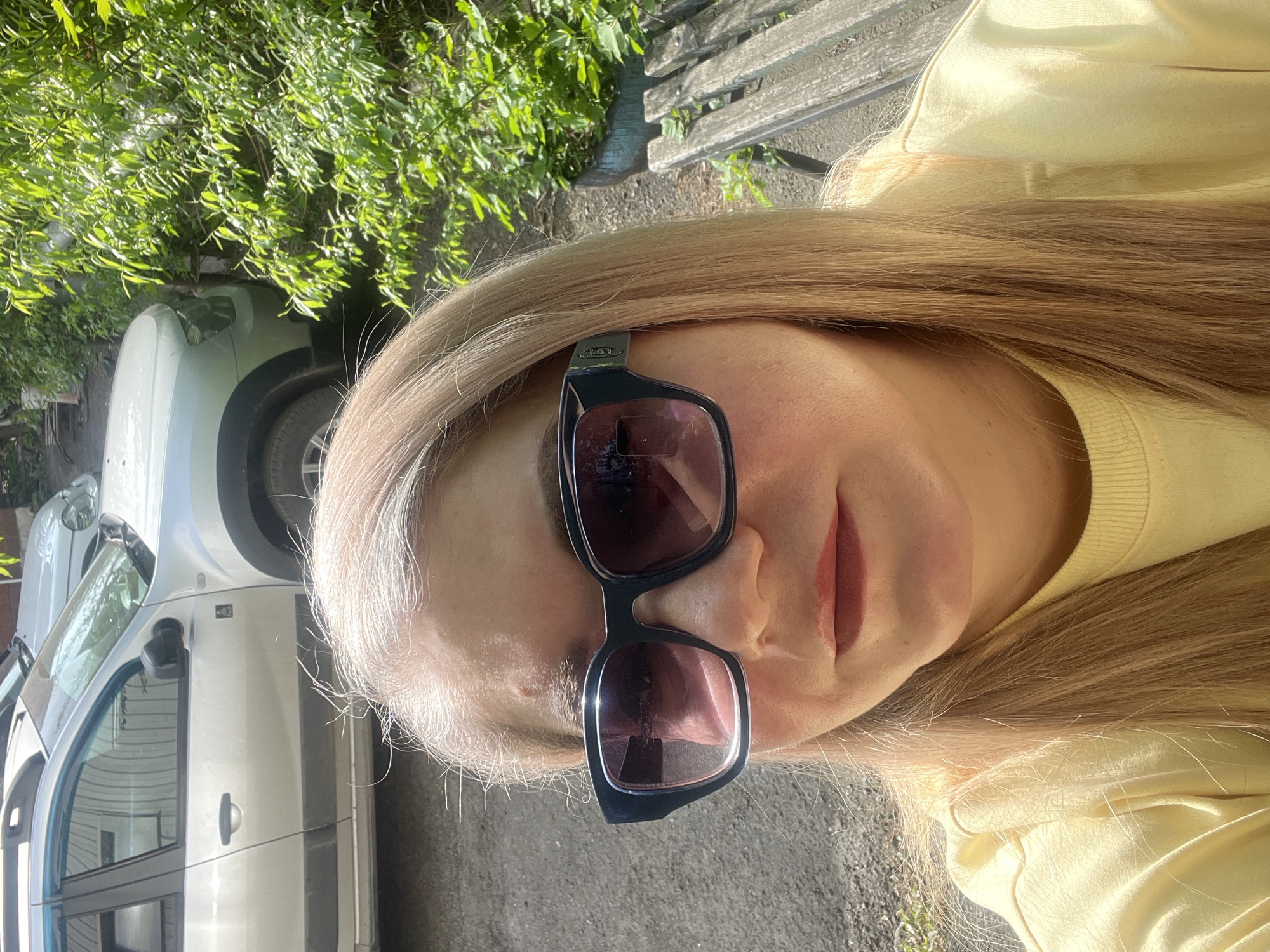A selfie taken outdoors on a sunny day. The subject is a fair-skinned person with long, straight blonde hair, wearing sunglasses and a yellow sweater. A white car is visible in the background, parked near a tree with dense green foliage. Part of a wooden bench is also visible. The location is potentially Izhevsk, Russia, but there are no discernible landmarks to definitively confirm.