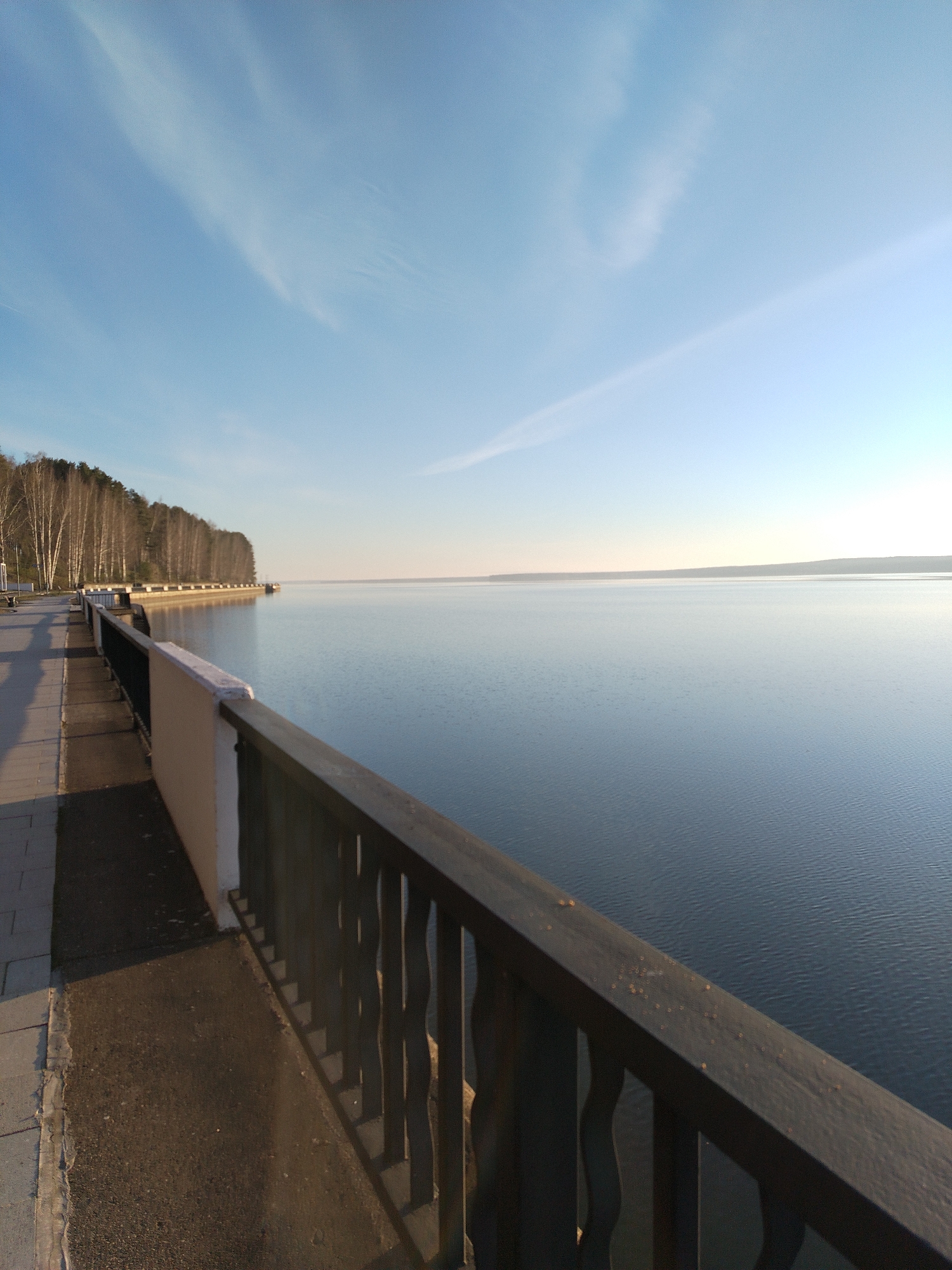 The image depicts a tranquil waterscape in Chaikovsky, Russia. A wooden railing lines a paved walkway that runs alongside a body of water, possibly a lake or reservoir. A row of trees extends along the left side, meeting the edge of the water. The sky is clear with faint wisps of clouds. The water surface is calm, reflecting the sky. The overall lighting suggests it is either early morning or late afternoon.