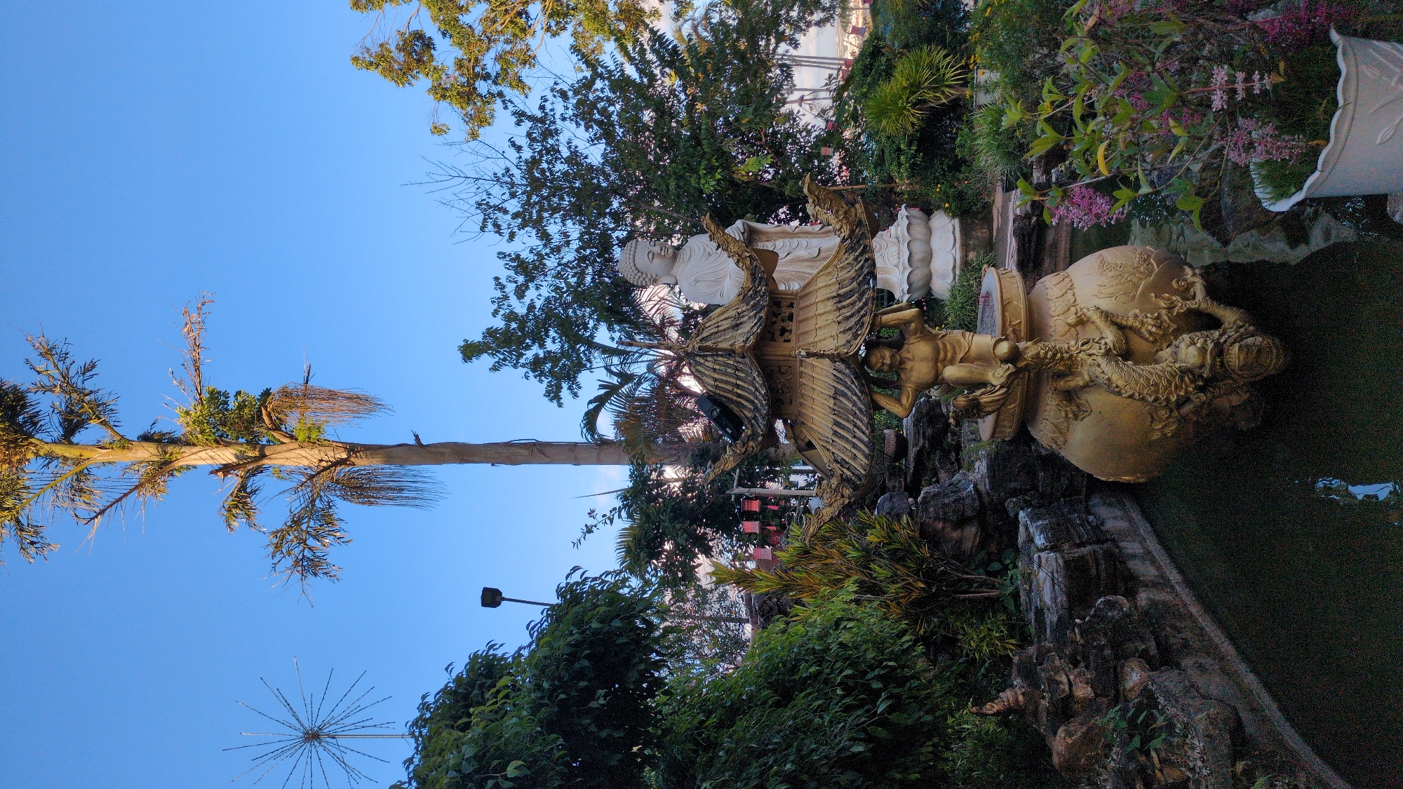 In a garden setting, possibly in Lâm Viên - Đà Lạt, Vietnam, a tall, slender tree with sparse foliage dominates the upper center of the frame, set against a clear blue sky. To the left of this tree, a starburst-shaped decorative light fixture is visible above dense green foliage.
In the mid-ground, a large, golden urn-like structure with intricate dragon carvings is a central feature. Perched atop this urn is a small, golden statue of a figure holding up a tiered, pagoda-like roof. Behind this golden structure, a large, white statue of Buddha, seated on a lotus base, stands serenely amidst lush greenery.
The foreground features a body of dark water, possibly a pond or water feature, with reflections visible on its surface. Rocky outcrops and various plants, including some with pink flowers in a white pot, surround the water and the statues. The overall scene suggests a tranquil, decorative garden with Buddhist-inspired elements.