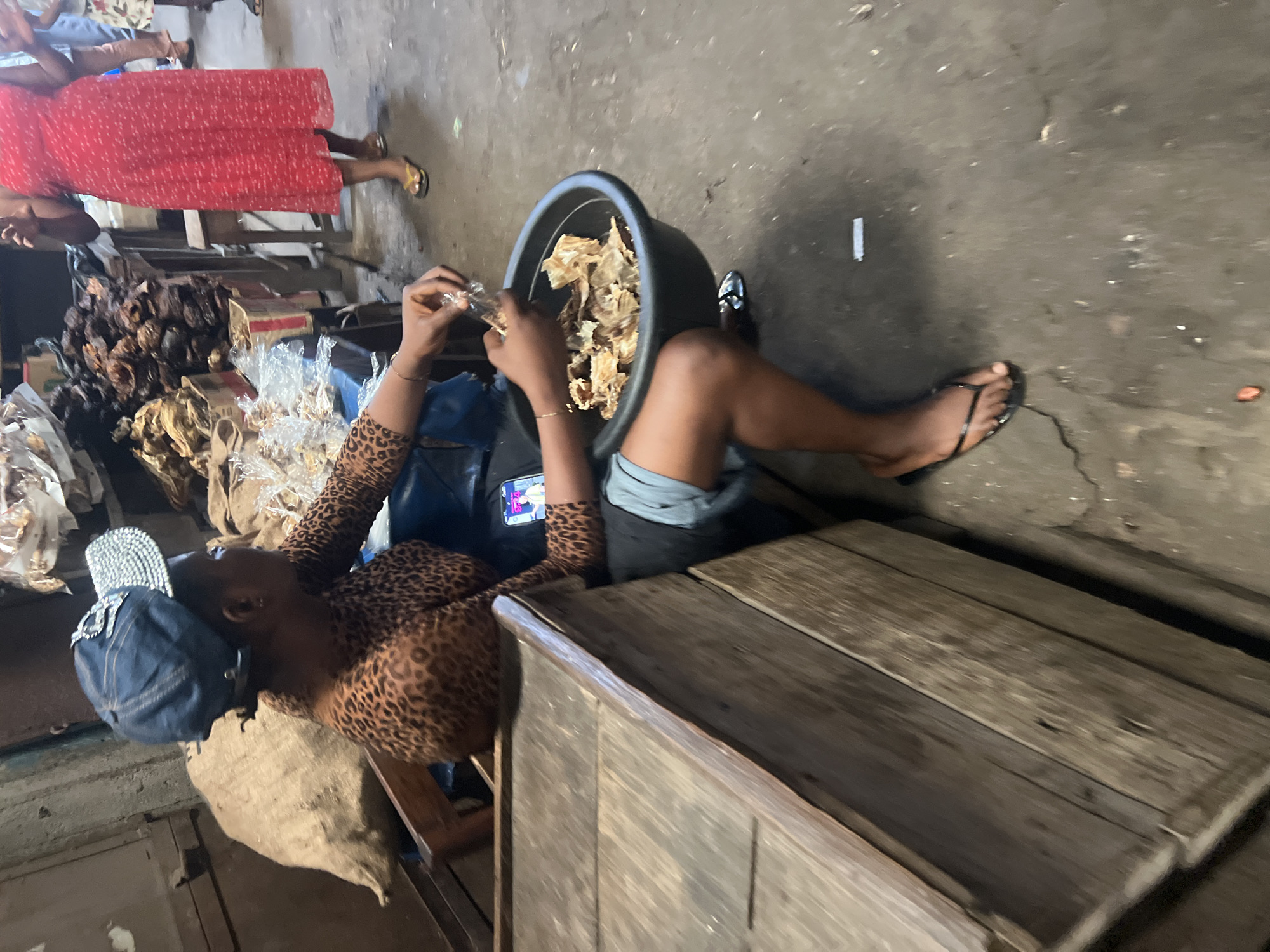 The image captures a scene in an indoor or covered market in Nkpor, Nigeria, likely during the daytime. The central subject is a woman who is lying or reclining on the concrete floor. She wears a leopard-print long-sleeved top, grey shorts, and black flip-flops, with a dark denim cap featuring metallic embellishments on her head. She is actively engaged in her work, holding a clear plastic bag over a large black basin filled with what appears to be dried food items, possibly dried fish or similar provisions. A mobile phone with an illuminated screen is tucked into her waist.
To the left of the woman, a substantial pile of dark, dried, and possibly smoked food products, resembling fish or meat, is visible on a wooden surface or stall. Various other goods are packaged in clear plastic bags nearby. In the upper left background, another person is partially visible, wearing a red patterned dress and gold flip-flops. The ground is a worn concrete surface, and to the right, a structure made of wooden planks suggests a market stall or counter. The scene depicts a typical, busy market environment with vendors attending to their goods. No legible text is visible in the image.