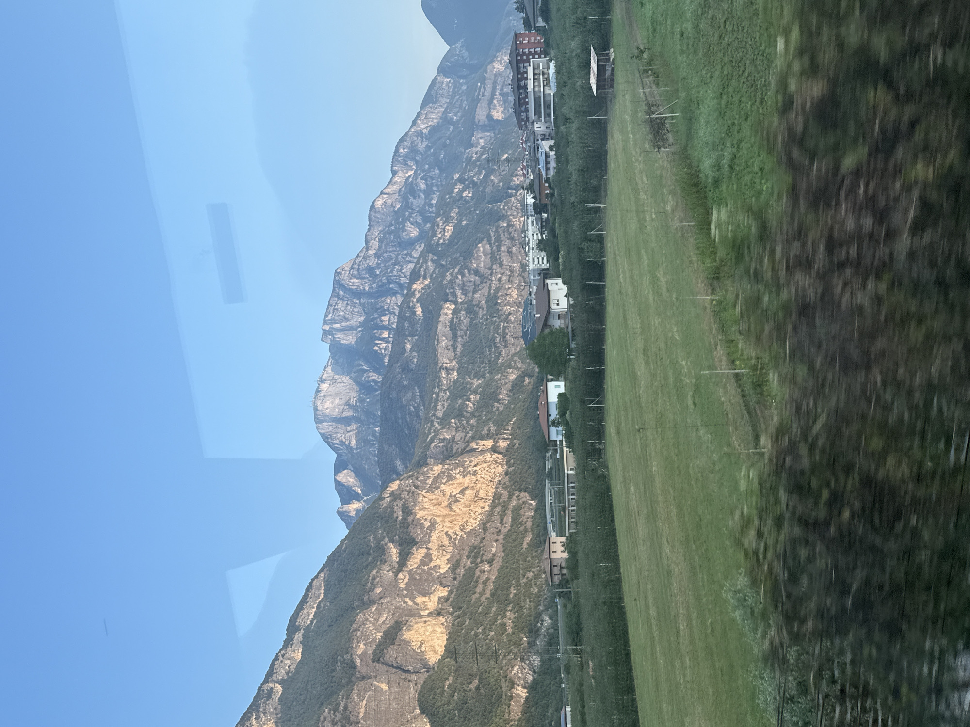 The image captures a scenic view of a valley with imposing mountains in the background, a settlement of buildings, and agricultural fields in the foreground. The mountains, bathed in the warm light of what appears to be late afternoon or early morning, display rugged terrain with patches of vegetation. A town or village is nestled at the base of the mountains, characterized by a cluster of residential buildings and some commercial structures. The foreground is dominated by a lush green field, possibly a farm or park, with some trees and fences visible. The sky is clear blue, with a hint of reflection from glass, suggesting the photo was taken from inside a vehicle or building. The overall impression is one of a serene, rural landscape adjacent to a built-up area. Based on the distinctive mountain formations and the proximity to a settlement, this scene is likely located in the vicinity of Trento, Italy.