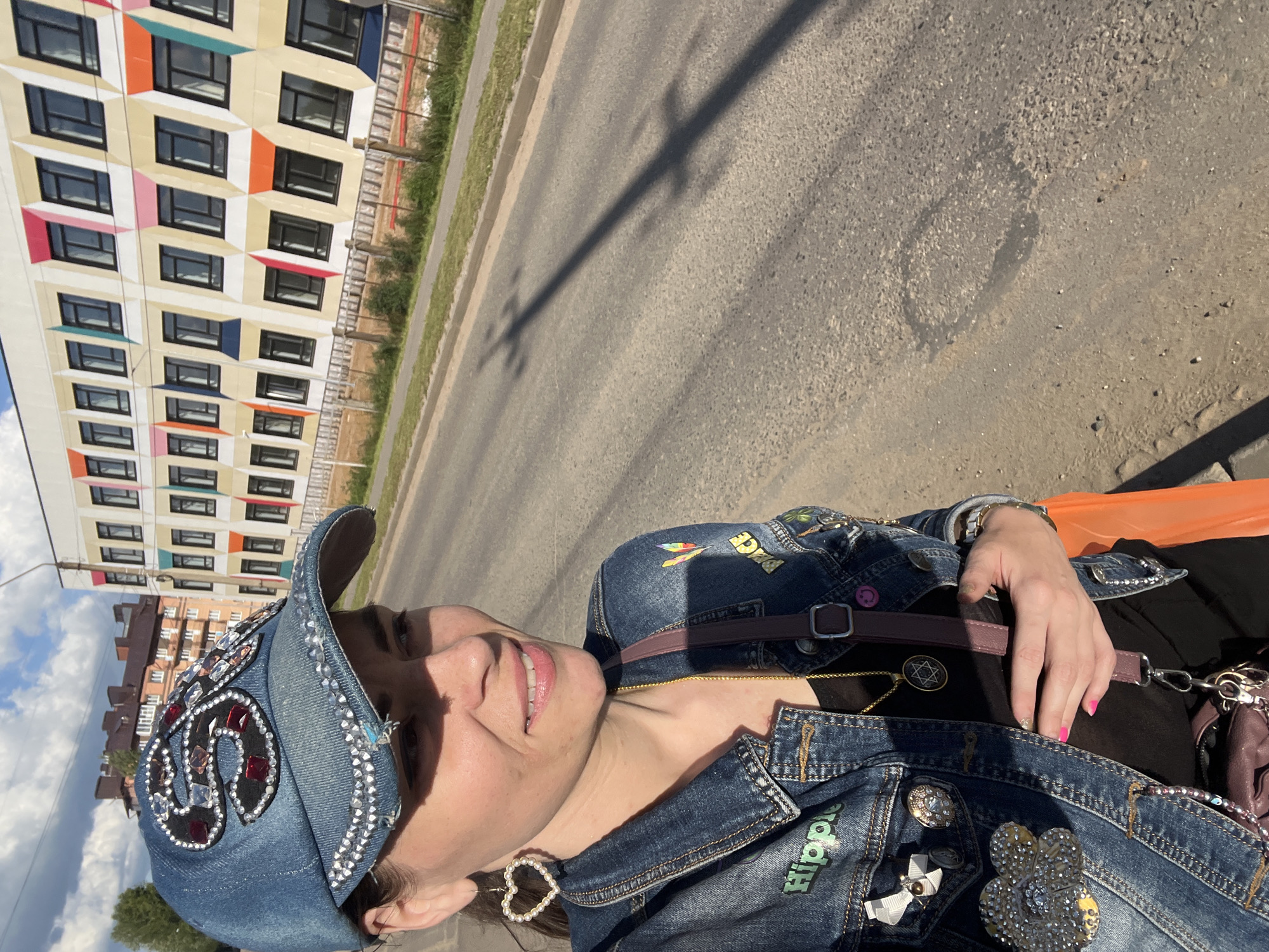 In this close-up, vertical outdoor shot, a woman is captured from the chest up, facing right and looking towards the camera with a slight smile. She wears a denim jacket adorned with patches, rhinestones, and buttons, including one that says "Hippie." A matching denim baseball cap, also embellished with rhinestones, sits on her head. A delicate heart-shaped pearl earring is visible on her left ear. She has a thin gold chain with a Star of David pendant around her neck.
Behind her, a brightly colored, modern building with a grid of windows stands to the left. The building's facade features blocks of yellow, orange, pink, and teal, creating a playful geometric pattern. To the right of the building, a paved road with a grassy verge stretches into the distance. The scene suggests it is daytime, with bright sunlight casting shadows, including a distinct shadow of what appears to be a utility pole across the road. The sky is visible in the upper left corner, with scattered clouds. The overall impression is a casual, sunny day, possibly in a suburban or semi-urban setting in Krasnoe-na-Volge, Russia, given the context.