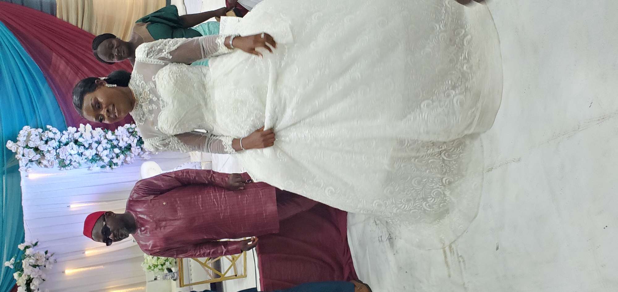 The image shows a bride in a white wedding dress, standing in what appears to be a wedding reception hall in Bukuru, Nigeria. She's holding up the front of her dress. Behind her is a man in a red suit with a matching cap and sunglasses, likely an elder or family member. There is a woman in a green dress behind the bride. The background includes floral decorations and draped fabric in shades of blue and burgundy. The floor is a light, reflective surface.