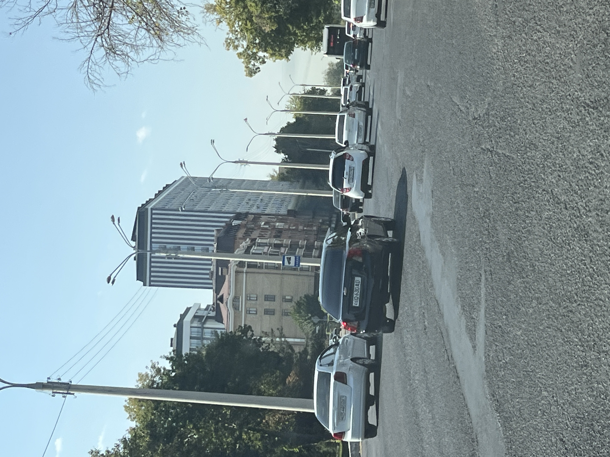 A traffic scene on a sunny day in Tashkent, Uzbekistan, shows a multi-lane road with several cars driving. In the foreground, a black Chevrolet Nexia is visible, followed by a white Chevrolet Lacetti. Further ahead, a convoy of white cars, including what appears to be a Daewoo Gentra, is moving. In the background, tall buildings with modern architecture, including a building with vertical stripes, line the street. Trees and streetlights are also present. The road surface shows some signs of wear and tear, with a distinct painted line indicating a lane change. A bus is seen in the distance, and a blue traffic sign is mounted on a pole. The sky is clear and blue.