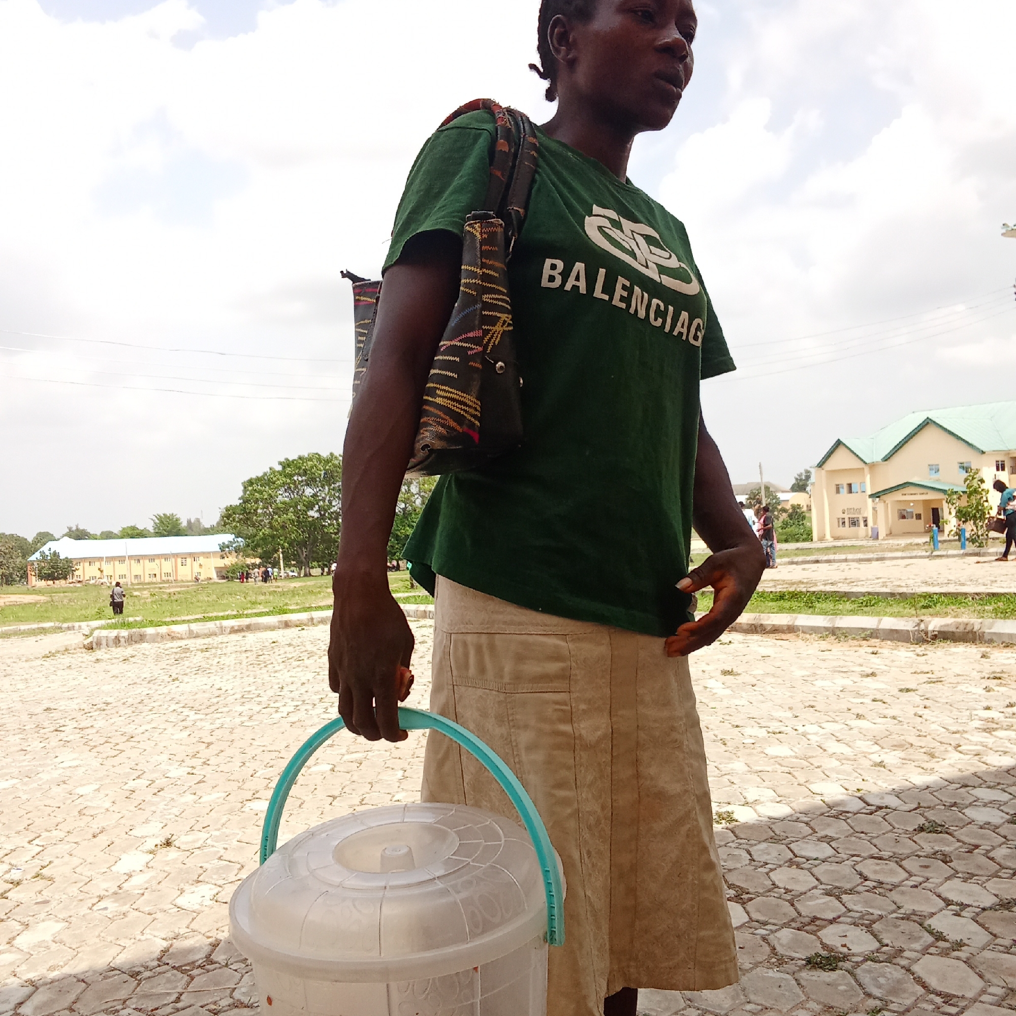 An eye-level outdoor shot depicts a woman standing in Akwanga, Nigeria, under a bright, partly cloudy sky. The woman, with dark skin and short, braided hair, is seen from the waist up, facing towards the right and looking slightly upwards. She wears a dark green t-shirt with a light-colored, stylized logo and the text "BALENCIAG" (likely Balenciaga) across the chest. A dark shoulder bag with colorful patterns hangs from her left shoulder.
She is dressed in a light tan or beige skirt. In her left hand, she holds a clear plastic bucket with a light blue handle. Her right hand rests casually on her side, touching her skirt.
The woman stands on a paved surface made of light-colored, hexagonal interlocking blocks. The background features a grassy area with scattered trees and several buildings. To the left, a long, light-colored building with a light blue roof is visible. To the right, another light-colored building with a green roof is seen, appearing to be a multi-story structure. Several other people are visible in the distance, though they are indistinct. The scene suggests everyday activity in a public or communal outdoor space during daylight hours.