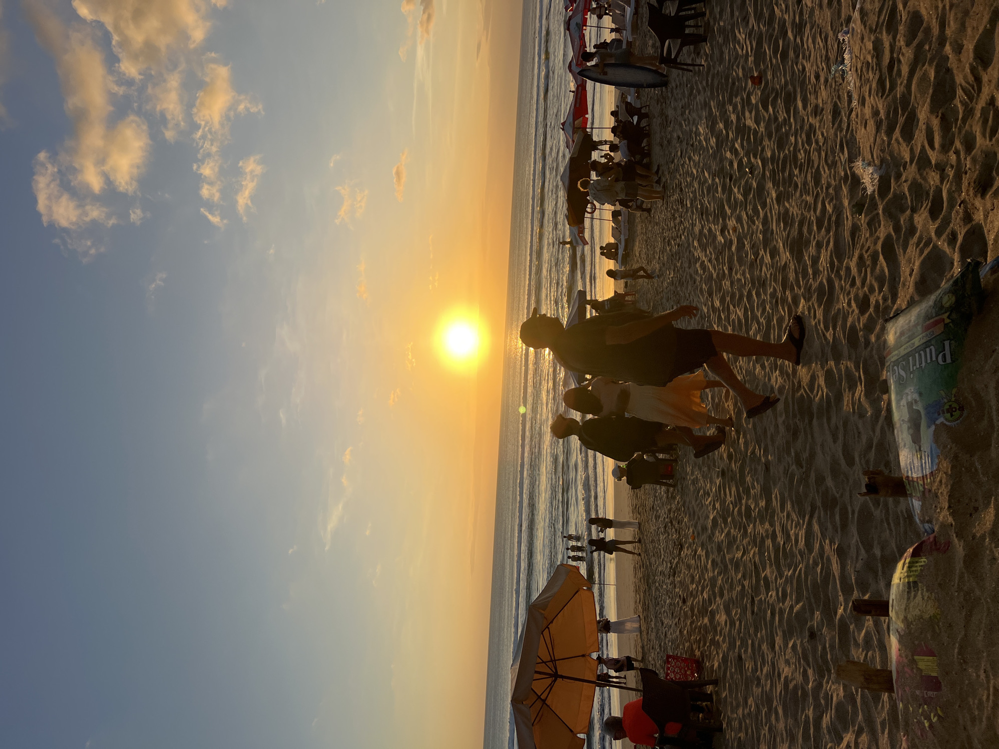 This is a photograph taken at sunset on Kuta Beach in Kuta, Indonesia. The sun, a bright orange orb, is low on the horizon, casting a warm glow across the sky and the ocean. The sky transitions from a soft blue at the top to shades of orange and yellow near the horizon, with scattered clouds tinged by the sunset.
The beach is sandy, with visible footprints and ripples in the sand. In the foreground, partially obscured by the sand, are some discarded items, including what appears to be a plastic bag with writing in a foreign language and some wooden sticks.
In the middle ground, several people are visible on the beach. Some are walking, while others are standing or sitting. To the left, an orange beach umbrella provides shade for a seated person. Further back, people are interacting in small groups, and some are wading in the gentle waves of the ocean. Several beach chairs and umbrellas are set up along the shoreline.
In the foreground, a group of people are silhouetted against the setting sun. A man in a dark shirt and cap is walking towards the viewer, while another person in a light-colored dress is walking away. To the right, a person with a surfboard is standing.
The overall scene depicts a lively beach atmosphere during a beautiful sunset. The lighting is soft and warm, characteristic of the golden hour.