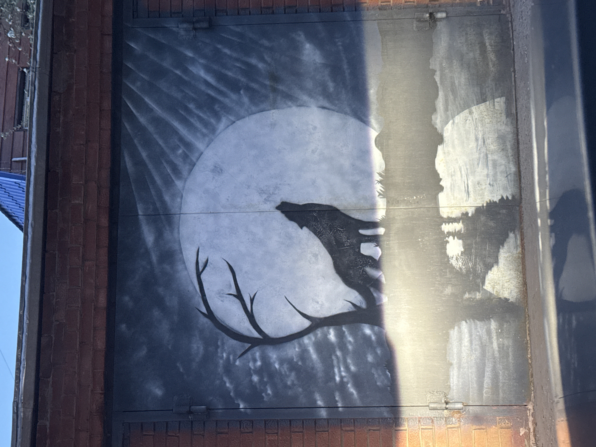 The image shows a mural painted on a metal door set into a brick wall. The mural depicts a wolf howling at a full moon, with a bare tree branch silhouetted in the foreground. The right side of the door has a faded painting of a forest landscape. The mural appears to be located on the exterior of a building, possibly in an urban area. The specific location is Votkinsk, Russia, based on provided context.