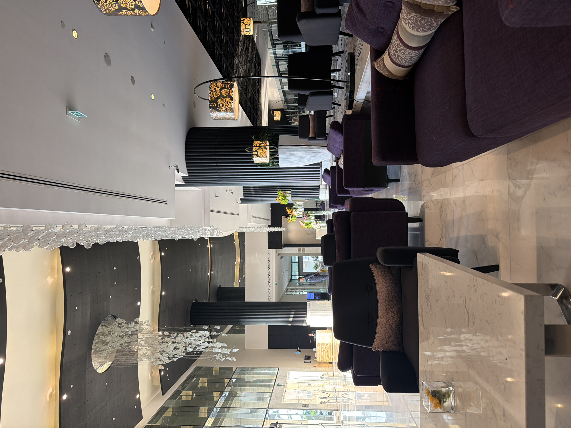 The image depicts the opulent interior of the lobby of the St. Regis Abu Dhabi, located on Corniche Road in Abu Dhabi, United Arab Emirates. The scene is characterized by a high-end, contemporary design with a focus on luxury and comfort.
In the foreground, a modern, white marble coffee table with sleek metallic legs is visible, holding a small glass vase with yellow flowers. Behind the table, plush, deep purple sofas and armchairs are arranged to create seating areas. These furniture pieces feature decorative throw pillows with intricate patterns in shades of beige and brown.
The background reveals a grand and expansive lobby. A striking feature is the elaborate crystal chandelier suspended from a dark, multi-tiered ceiling, creating a dramatic focal point. Tall, fluted black columns punctuate the space, adding architectural interest and a sense of scale. Large windows and glass doors offer glimpses of the exterior and allow natural light to flood the area.
In the middle ground, a reception desk with a distinctive gold-toned surface is manned by a person in a uniform. Further back, another individual in a suit is standing near a display screen, likely a hotel staff member.
The flooring is a polished white marble, reflecting the ambient light and contributing to the overall sense of sophistication. The lighting is a mix of recessed ceiling lights and decorative lamps with patterned shades, casting a warm glow throughout the lobby. The overall atmosphere is one of refined elegance and spaciousness.
