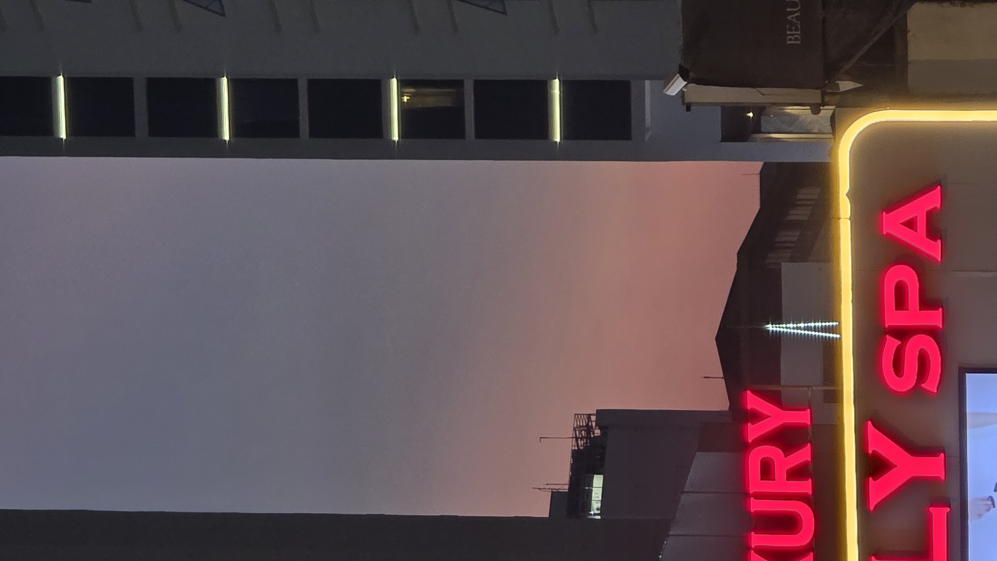 The image shows a twilight urban scene in Nha Trang, Vietnam. The foreground contains a tall building with a facade of regularly spaced dark windows and illuminated vertical elements. Adjacent to this building is a vertical neon sign reading "LUXURY SPA," with the letters in pink outlined by yellow neon. The sky in the background exhibits a gradient, shifting from a darker purplish-gray at the bottom to a lighter pinkish-orange towards the upper-middle. Silhouettes of other buildings and structures are visible against the sky, including what appears to be a communication tower with blinking lights. A sign with the partial word "BEAU" is visible. The overall impression is of a city at dusk with the illuminated signs and building features standing out against the fading light.