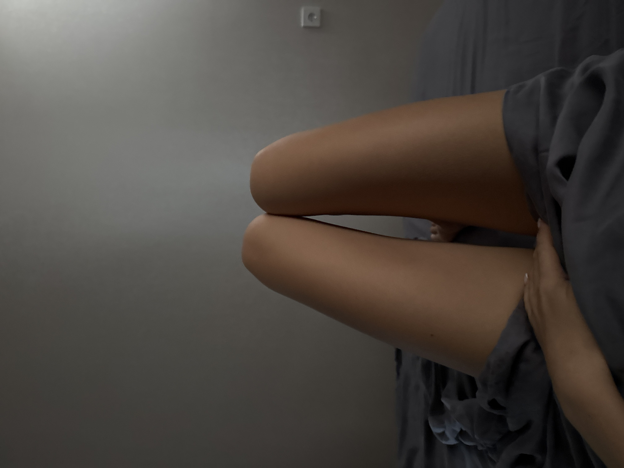 The image shows a close-up, low-angle view of a person's legs lying on a dark gray bed. The person is covered from the waist down with a dark gray blanket, and their knees are slightly bent and close together. The skin tone of the legs is light brown. The background is a plain, textured gray wall with a white electrical outlet on the right side. The lighting is dim, casting a shadow on the wall and creating a soft gradient. The overall impression is one of relaxation or rest in an indoor setting.