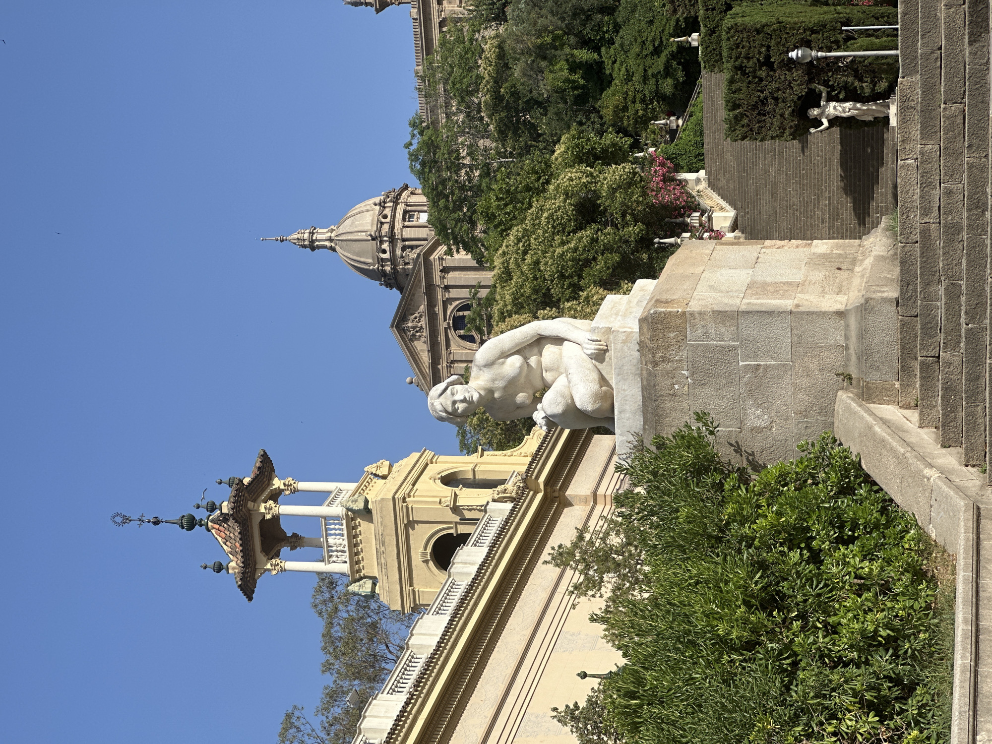 This is a photograph of the Palau Nacional, the National Palace of Catalonia, situated on Montjuïc hill in Barcelona, Spain. The image captures the iconic dome and grand architecture of the palace, which serves as the main building of the Museu Nacional d'Art de Catalunya (MNAC). In the foreground, a classical stone sculpture of a kneeling nude woman is prominently displayed on a pedestal. Behind the sculpture, a grand staircase ascends towards the palace, lined with manicured hedges and decorative urns. To the left of the staircase, another smaller classical sculpture of a male figure stands next to a lamppost, partially obscured by greenery. The sky is a clear, bright blue, indicating a sunny day. The overall scene depicts a grand, historic architectural site with classical art and landscaping.