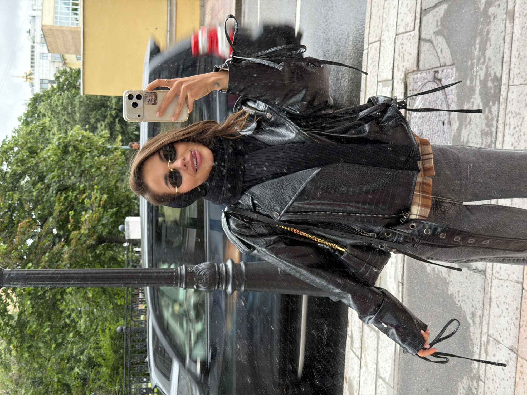 The image depicts a young woman taking a selfie on a city street, likely in Moscow, Russia, given the architectural style and the presence of a lamppost that is commonly seen in Russian cities. The woman is wearing a black leather jacket with a crocodile-like texture, a black scarf, and grey jeans. She has on oval-shaped sunglasses and is sticking her tongue out playfully. In her right hand, she holds a smartphone to take the picture. Behind her, a black car is blurred due to motion. The street appears wet, suggesting recent rain. The background shows trees and buildings, with one building having a yellow facade and distinctive architectural features.