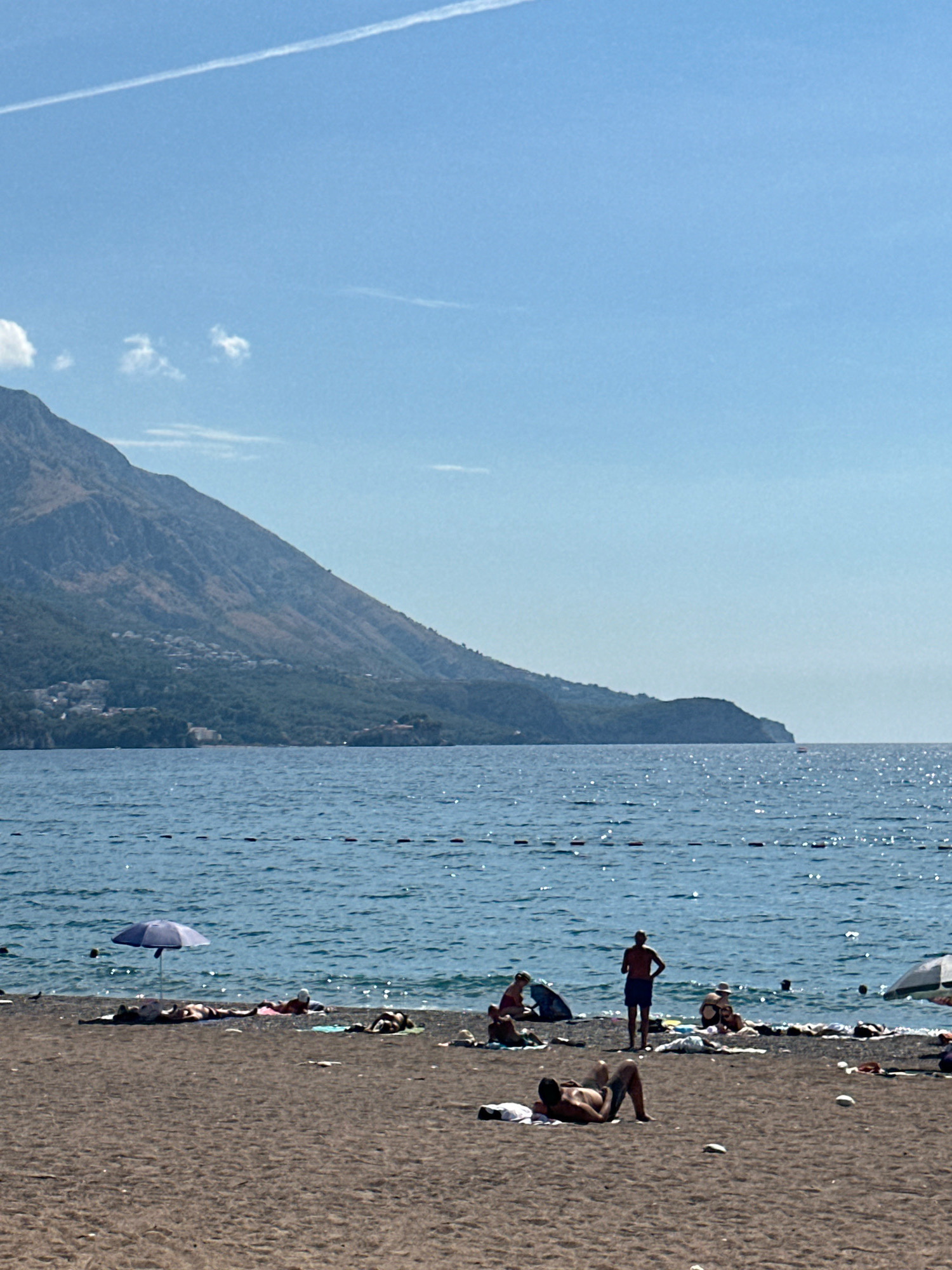 In Boreti, Montenegro, a sunny beach scene unfolds. The dark sand is dotted with several individuals sunbathing or relaxing. In the foreground, a man lies on his back on a white towel, his legs bent. Further back, an umbrella shades a few sunbathers. A man stands near the water's edge, facing away from the camera, wearing blue shorts. The sea, a deep blue, shimmers with sunlight, and a line of buoys marks a boundary in the water. In the distance, a lush green mountain slopes down to the coastline, with buildings nestled on its lower slopes. The sky is a clear, bright blue, with a faint white contrail stretching across it.