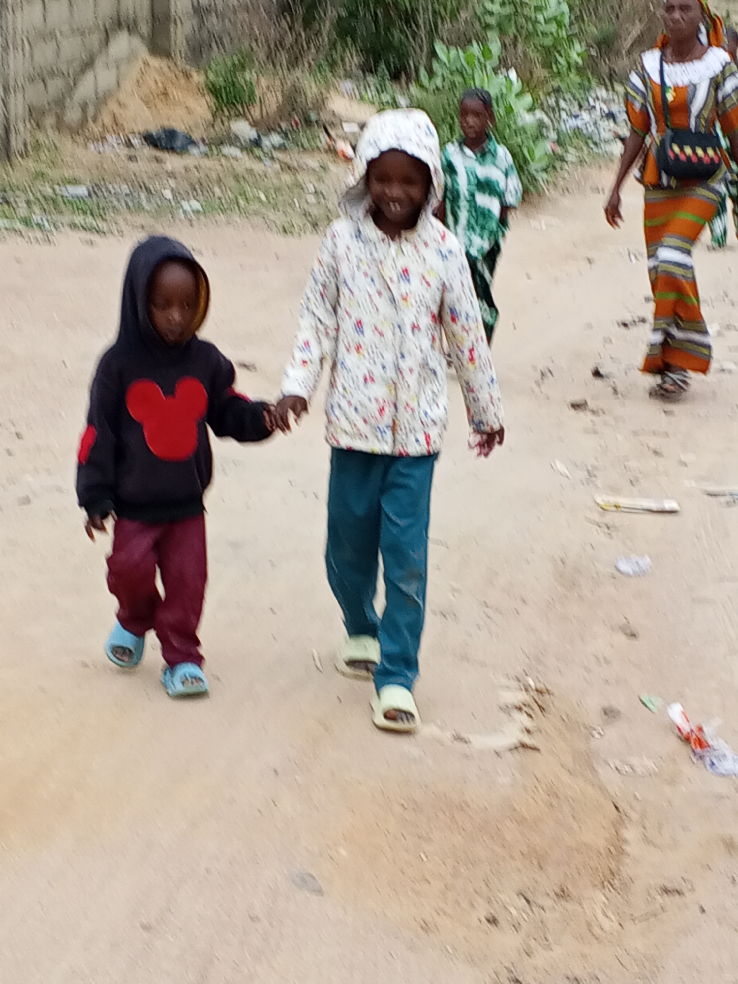The image shows two young children holding hands, walking on a dirt road in Adonkolo, Nigeria. The child on the left wears a dark hoodie with a red Mickey Mouse patch, maroon pants, and blue sandals. The child on the right wears a white patterned jacket with a hood, blue pants, and light green sandals. Behind them, two adults are walking in the same direction. The adult on the right is wearing a colorful orange and white patterned dress, and a black purse. The surrounding environment includes a partially constructed brick wall and scattered vegetation. There is litter on the ground.