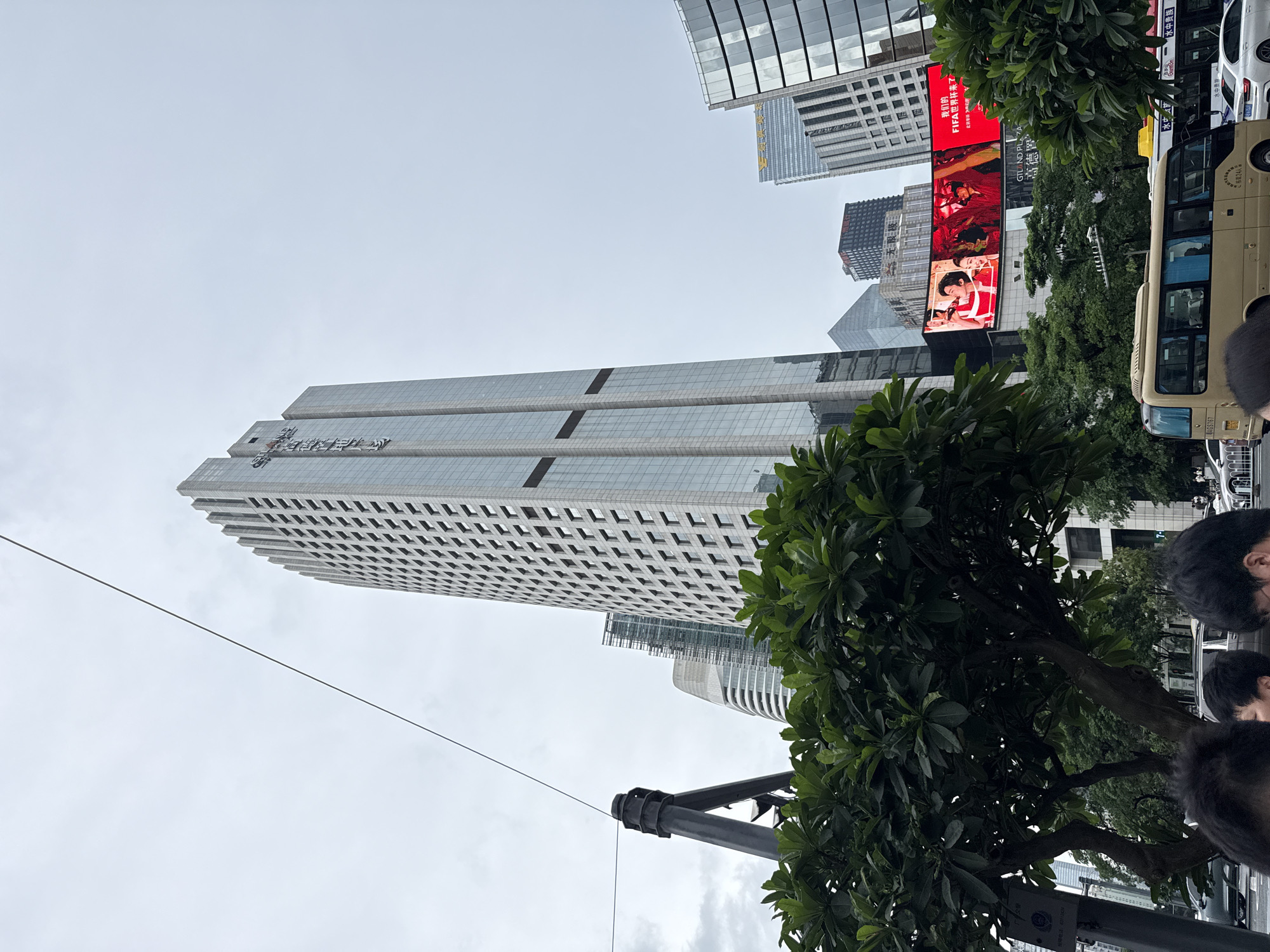 This image depicts a street scene in Tianhe District, China, dominated by towering modern skyscrapers. The perspective is from street level, looking upwards. A prominent feature is a large, glass-fronted office building with a grid-like facade. To the right, a beige bus is parked, with visible text on its side indicating its capacity ("板载24人") and a license plate number ("AGS767"). Above the bus, a large digital billboard displays vibrant advertisements, including a red banner with Chinese text, possibly related to the FIFA World Cup, and images of people.
The scene is set against an overcast sky, suggesting a cloudy day. Lush green trees are strategically placed throughout the urban landscape, adding a touch of nature amidst the concrete and glass. Several people are partially visible in the foreground and on the bus, their faces mostly obscured, giving a sense of everyday life in a bustling city. The presence of the digital billboard and the modern architecture points to a commercial or downtown area. Text visible includes "报障电话:63112424" (Fault reporting hotline), "无限极" (Infinitus, a health products company), "GTLAND PLAZA" and "高德置" (likely a shopping mall or commercial complex), and "水中贵族" (Aquatitude, a bottled water brand). The overall impression is one of a dynamic, urban environment in China.