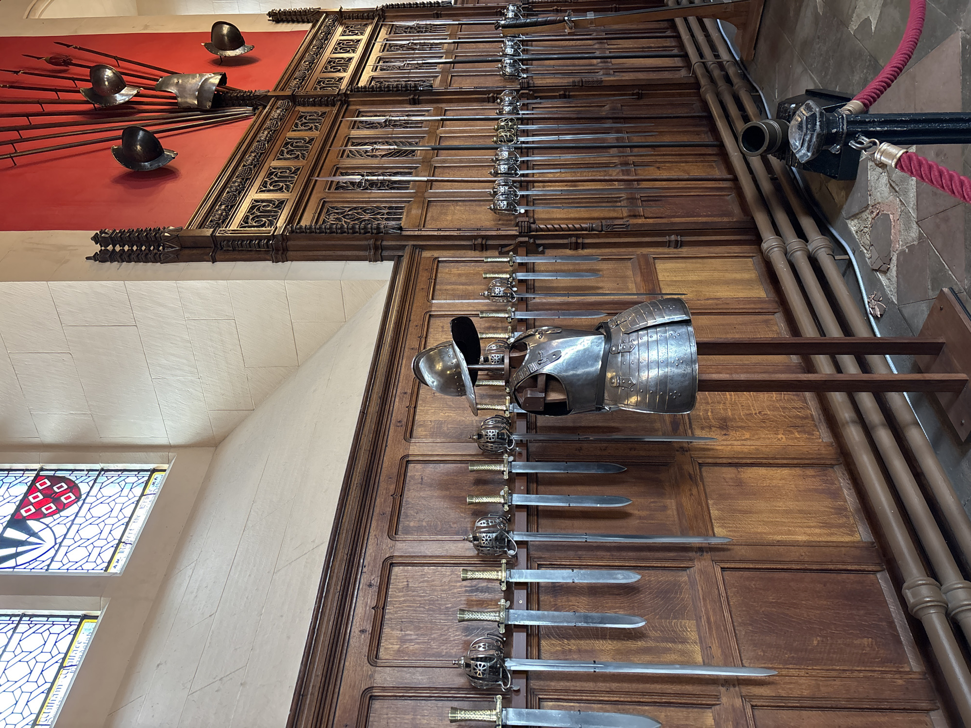The image shows a display of historical armor and weaponry, likely within a museum or historic building in the City of Edinburgh, United Kingdom. A collection of polearms (spears and halberds) is mounted on a red wall at the top left. Below them, suits of armor are mounted on a dark brown paneled wall with detailed carvings. A row of swords is arranged below the armor. Near the right edge of the image, there are vertical columns or pipes, indicating the wall structure and some security barrier, and a partial view of a window with stained glass is visible.