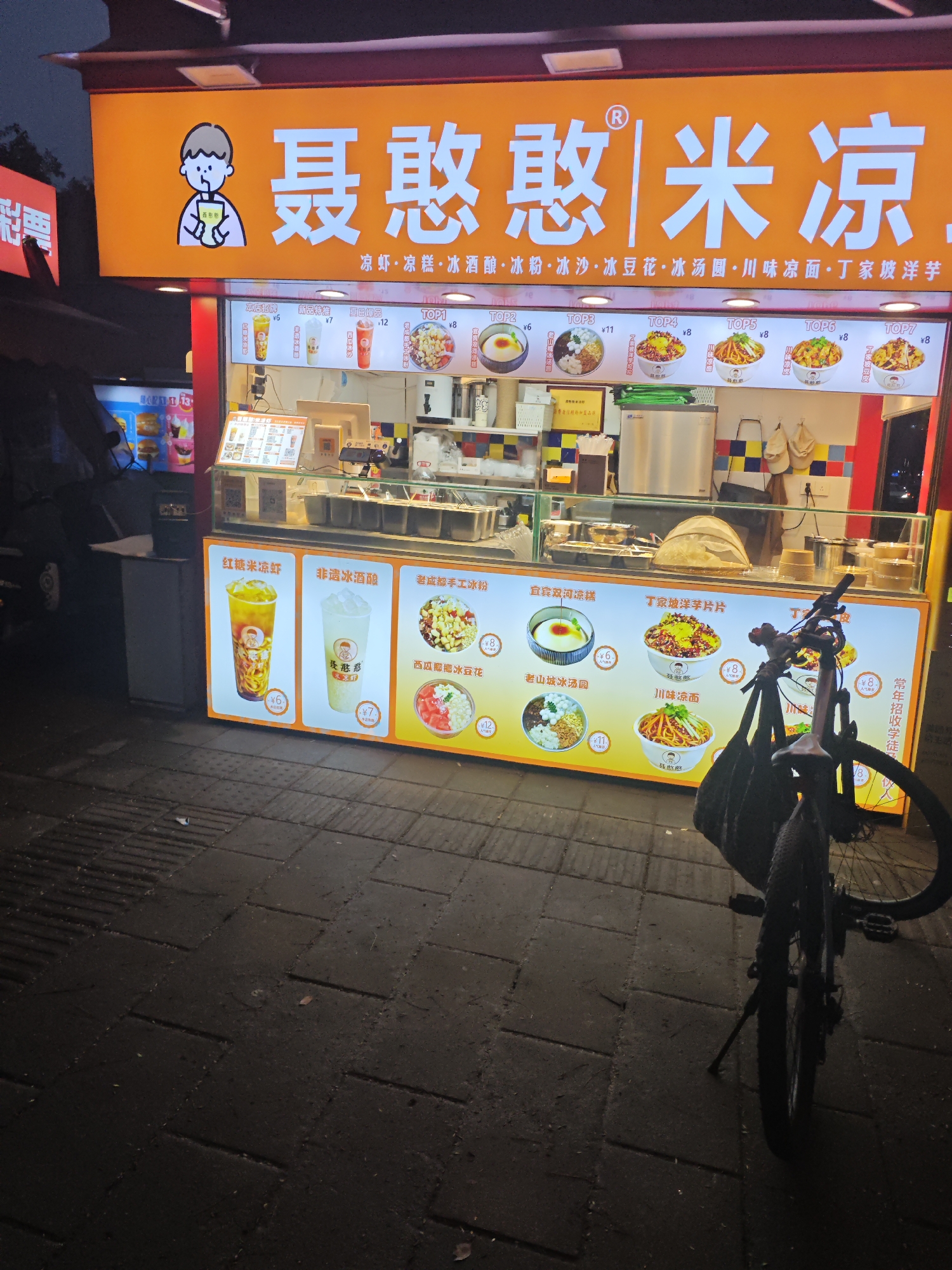Here is a detailed description of the image:
**Scene Overview:**
The image depicts a brightly lit food stall in what appears to be an outdoor market or street food area. The food stall is well-lit with an orange awning displaying the name "聂憨憨|米凉," which translates to "Nie Hanhan Rice Jelly," in large Chinese characters. The food stall is in Chongqing High-tech Zone, China. The storefront features a colorful cartoon character and advertisements for various cold desserts and noodle dishes. A bicycle is parked to the right of the food stall, partially obstructing the view.
**Subjects and Objects:**
* **Food Stall:** The central focus is the food stall, showcasing its offerings. It is brightly lit with a menu displayed on top, featuring photos of various dishes such as rice jelly, ice wine, ice powder, ice bean curd, ice dumplings, and Sichuan cold noodles.
* **Menu and Advertisements:** The front of the stall is adorned with illuminated menu boards showing images and prices of various dishes. The dishes include red sugar rice jelly, non-ice wine, old Chengdu handmade ice powder, Yibin Shuanghe Lianggao, and Dingjiapo yam slices.
* **Bicycle:** A black bicycle stands to the right of the stall, partly blocking the view of the menu board.
**Environment:**
* **Street/Market:** The food stall is located on a dark, paved street, suggesting an outdoor food area.
* **Lighting:** The scene is well-lit by the stall's bright orange sign and the illuminated menu boards. The background is darker, suggesting it is evening.
**Action:**
* **Commerce:** The food stall is actively open for business, ready to serve customers.
* **Parking:** Someone has parked a bicycle near the food stall, indicating potential customers or staff.
In summary, the image captures a vibrant food stall in an urban setting, highlighting its menu and promotional materials with a bicycle parked nearby.