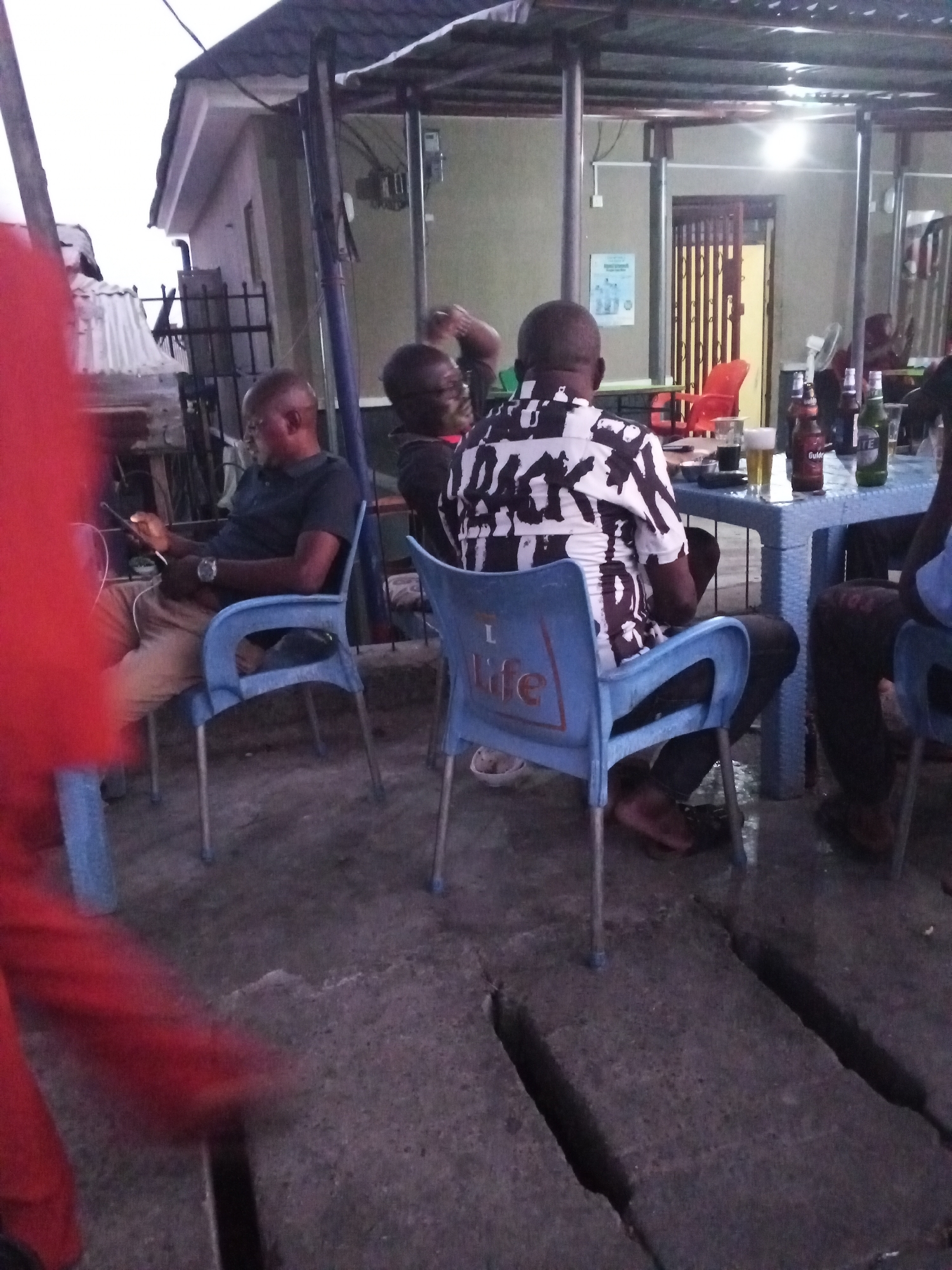 The image shows a gathering of people at an outdoor bar or restaurant in Lokoja, Nigeria. Three men are seated around a blue plastic table where bottled beverages (likely beer) and glasses are placed. The man in the foreground wears a black and white patterned shirt and sits in a blue plastic chair. Another man sits to his left, looking at a phone. The environment has a concrete floor with visible cracks, and a partially visible building is in the background. The lighting is dim, indicating it might be evening.