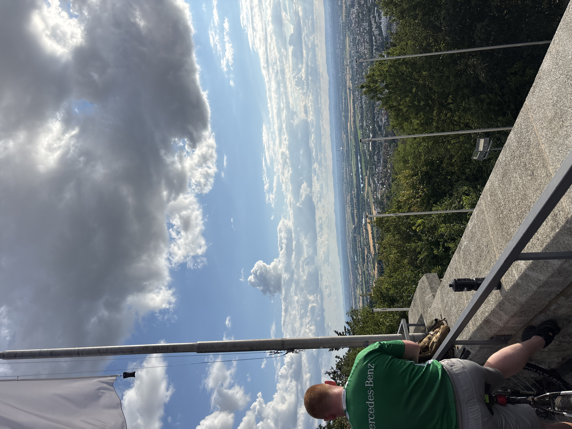 A person wearing a green "Mercedes-Benz" shirt and grey shorts sits on a bicycle on a stone wall overlooking a city. The person's back is to the camera. A large white flag on a pole is to the left of the frame. The sky is blue with large, white, and grey clouds. The city below is nestled among green trees. In the distance, there are mountains. The overall scene suggests a scenic overlook, possibly the Weinheimer Aussichtspunkt (Weinheim viewpoint).